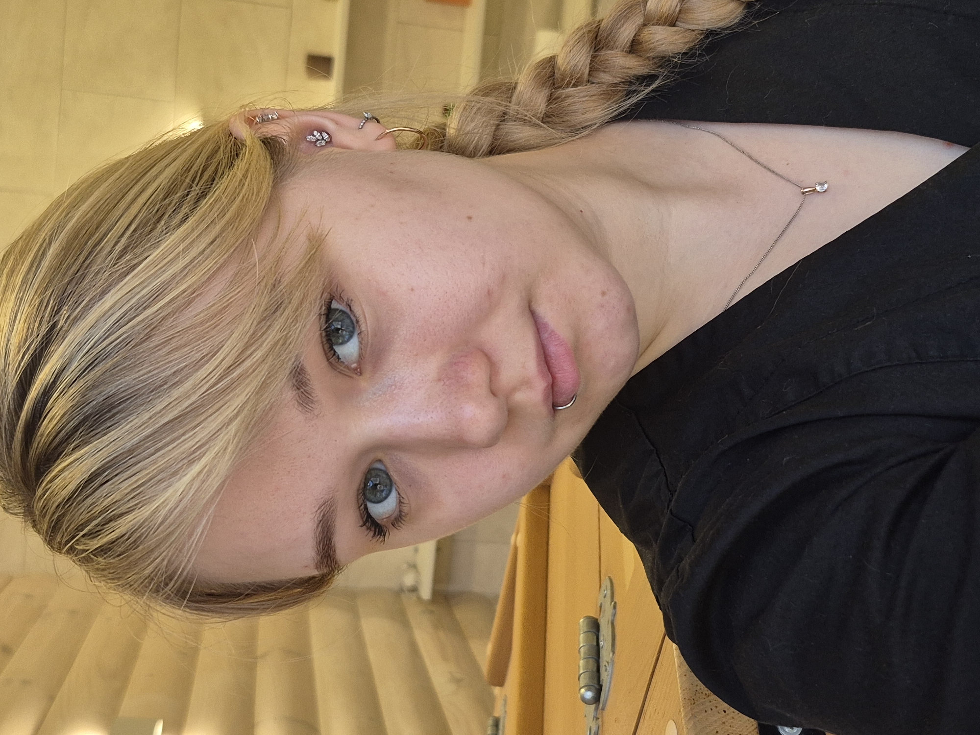 A fair-skinned female is positioned in a tilted close-up shot, displaying a braided hairstyle and blue eyes. Multiple piercings are visible in her left ear. A thin necklace with a small pendant adorns her neck, and a lip ring is present. She wears a dark shirt. The background suggests an interior setting with wooden elements like a door and flooring. There is no specific evidence that this image was taken in the city of Chaikovsky, Russia.