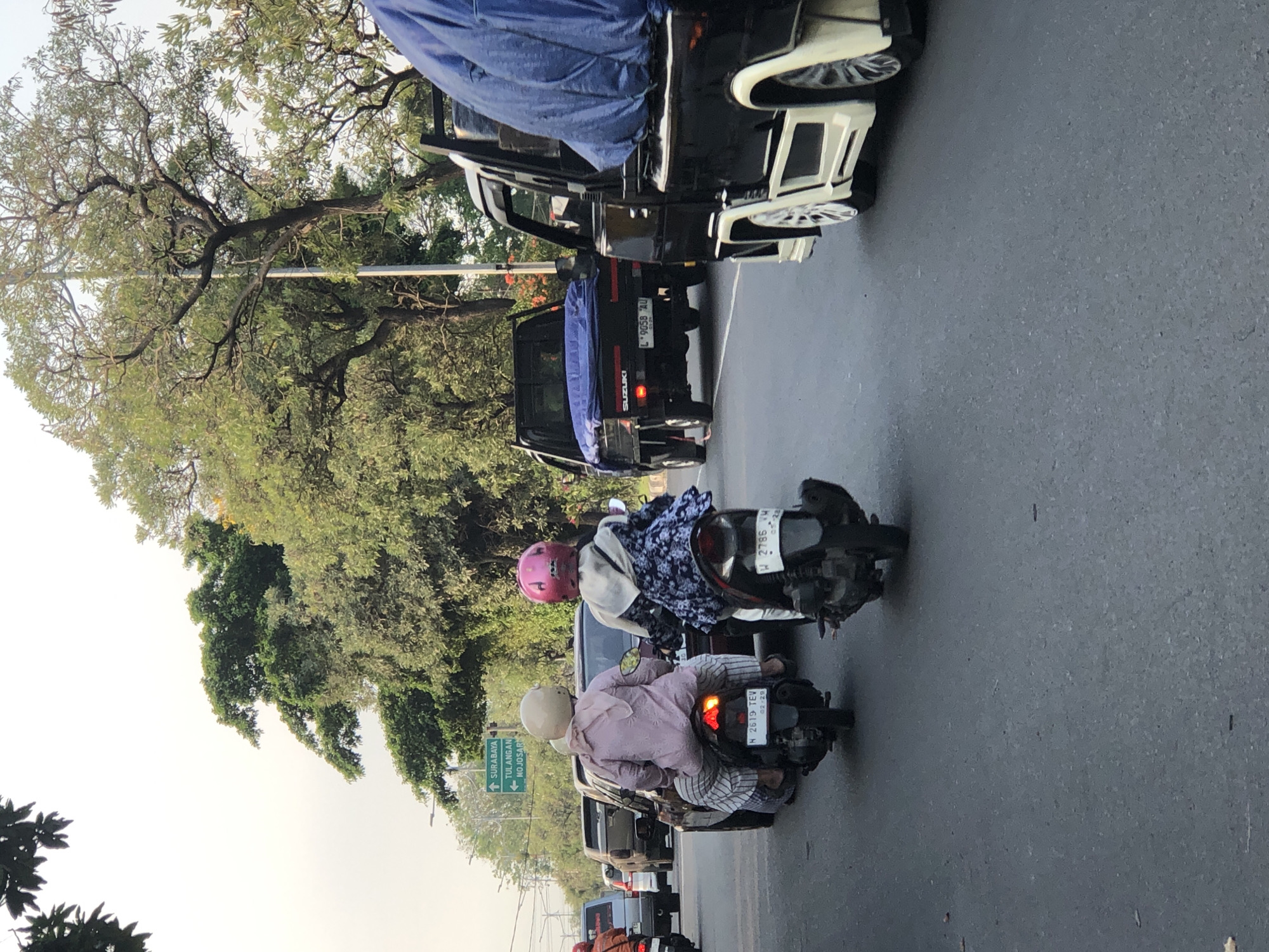 Two people are riding on a scooter in traffic on a street in Kludan, Indonesia. The person in front is wearing a pink hijab and a patterned blue and white long-sleeved shirt, and the person behind is wearing a beige hijab and a light pink long-sleeved shirt. They are both facing away from the camera. The scooter's license plate reads "N 2619 TEV". Other vehicles, including a black Suzuki pickup truck with a blue tarp on its bed and a white and black utility vehicle, are also visible in the traffic. A green street sign points to "SURABAYA" and "TULANGAN MOJOSARI". Lush green trees line the street. The sky is hazy and bright.