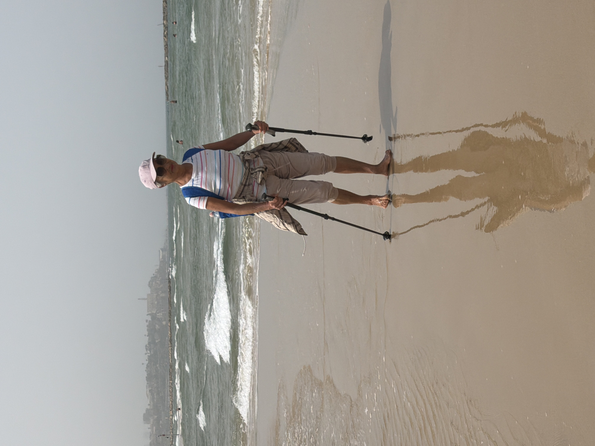 A man stands on a wet sandy beach in Tel Aviv, Occupied Palestine. He wears a pink baseball cap, a striped shirt, and khaki shorts. He holds two black trekking poles. A light-colored plaid shirt is tied around his waist. He is barefoot, and his shadow stretches out on the wet sand. Behind him are waves and a faint outline of a city skyline.