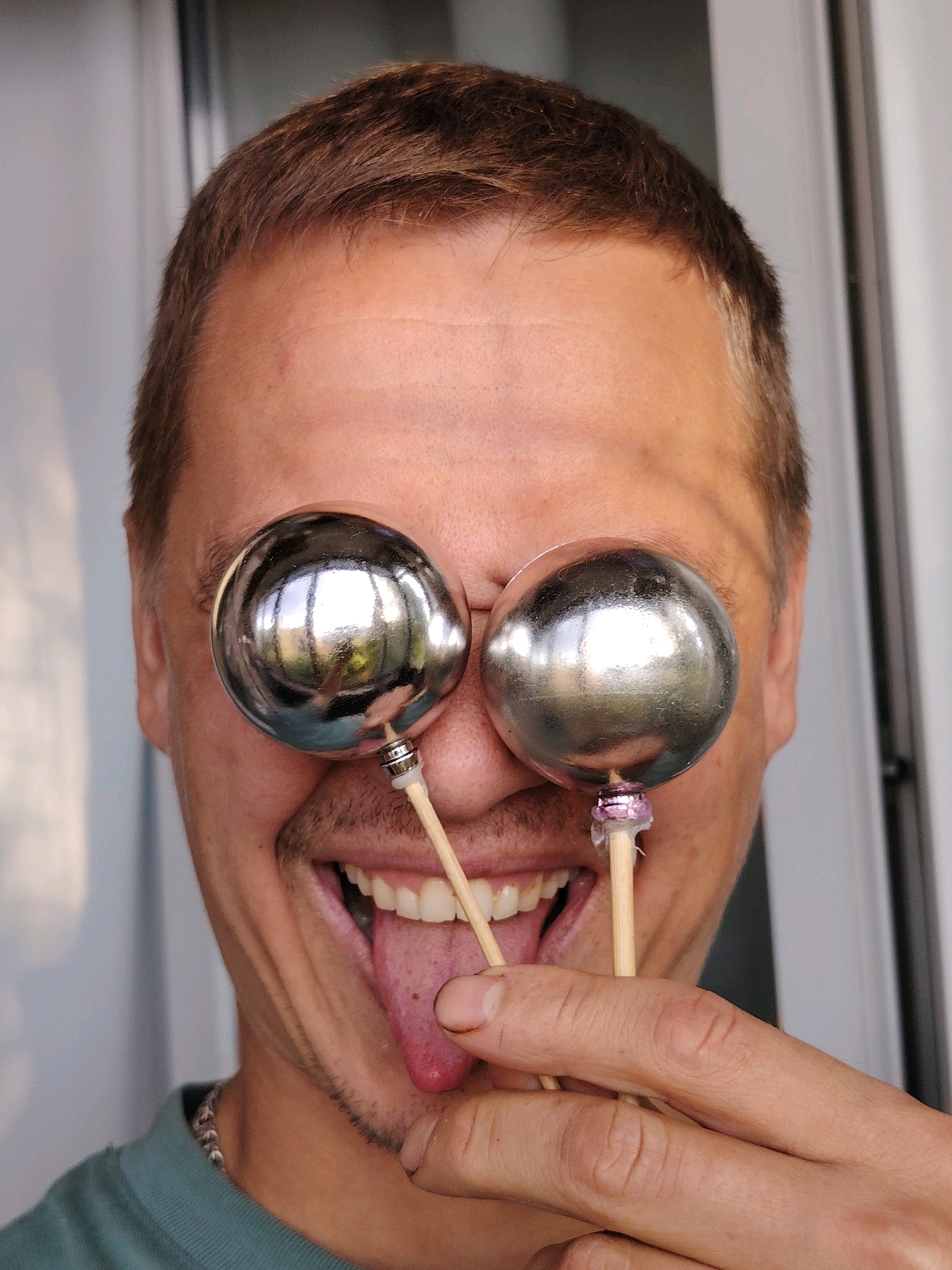 The image is a medium shot of a man in Chaikovsky, Russia, holding two shiny, spherical objects to his eyes like oversized novelty glasses. The man's mouth is open, with his tongue sticking out, and he is looking directly at the camera. His hair is short and brown. The background appears to be an indoor setting with a blurred light-colored wall and a vertical element, possibly a door or window frame. The man is wearing a teal-colored shirt and a beaded necklace.