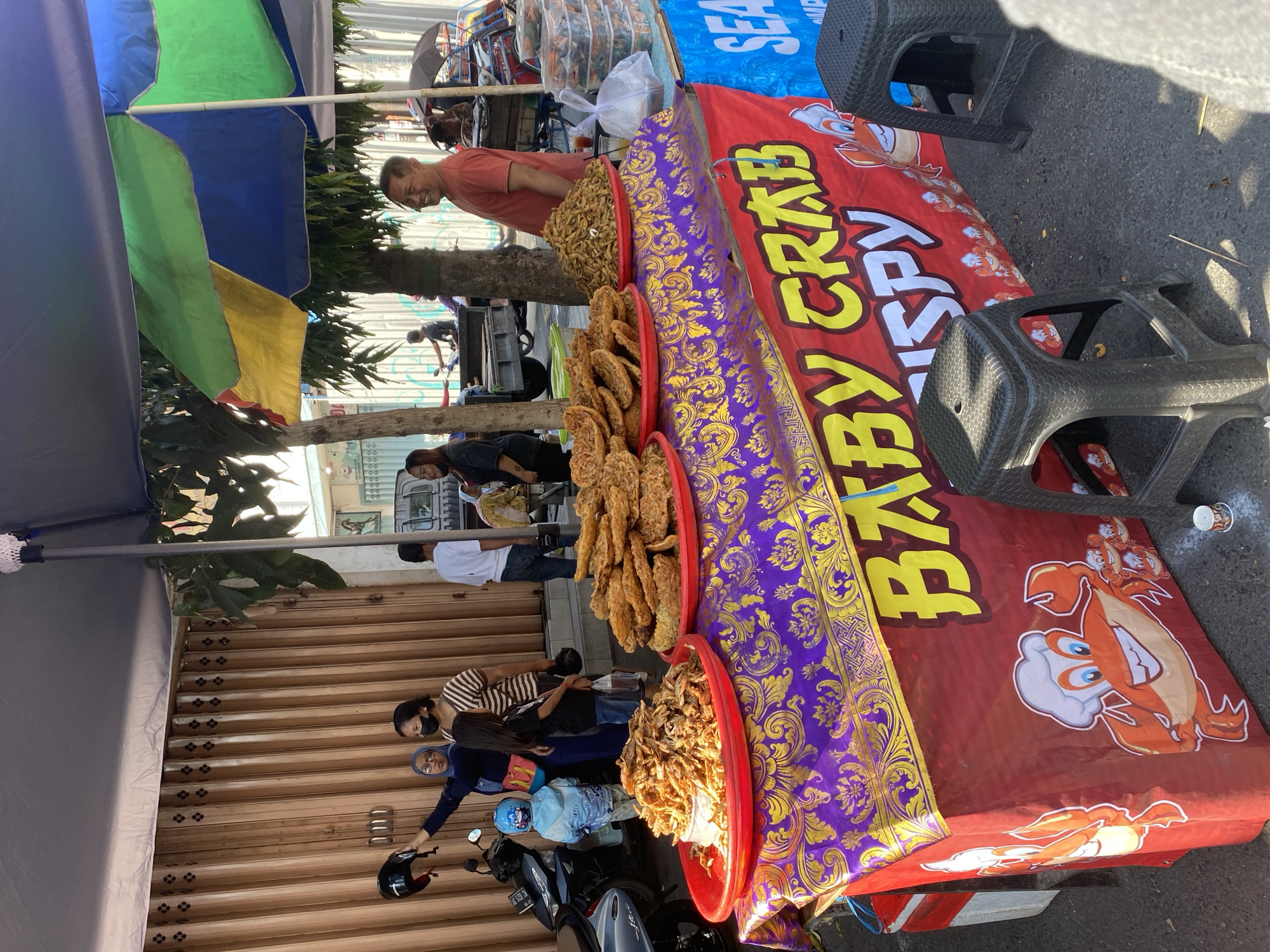 In Kediri City, Indonesia, on a street, a food stall is set up under a blue and green awning. The stall features a red cloth tablecloth with a purple and gold patterned runner, displaying various fried snacks. Two red platters are piled high with these fried items, one appearing to be small fried fish and the other larger, flatter fried fritters. Behind the food, a stack of clear plastic containers holds more of the fried snacks. A banner on the front of the stall reads "BABY CRAB CRISPY" and features cartoon illustrations of crabs, one wearing a chef's hat. Two black plastic stools are placed in front of the stall. Several people are visible, including a woman in a blue headscarf holding a black helmet, a woman wearing a face mask and a striped shirt, and a child in a light blue jacket. A man wearing an orange t-shirt is standing to the right, smiling at the camera. In the background, there's a corrugated metal fence, a parked truck, and other pedestrians.
