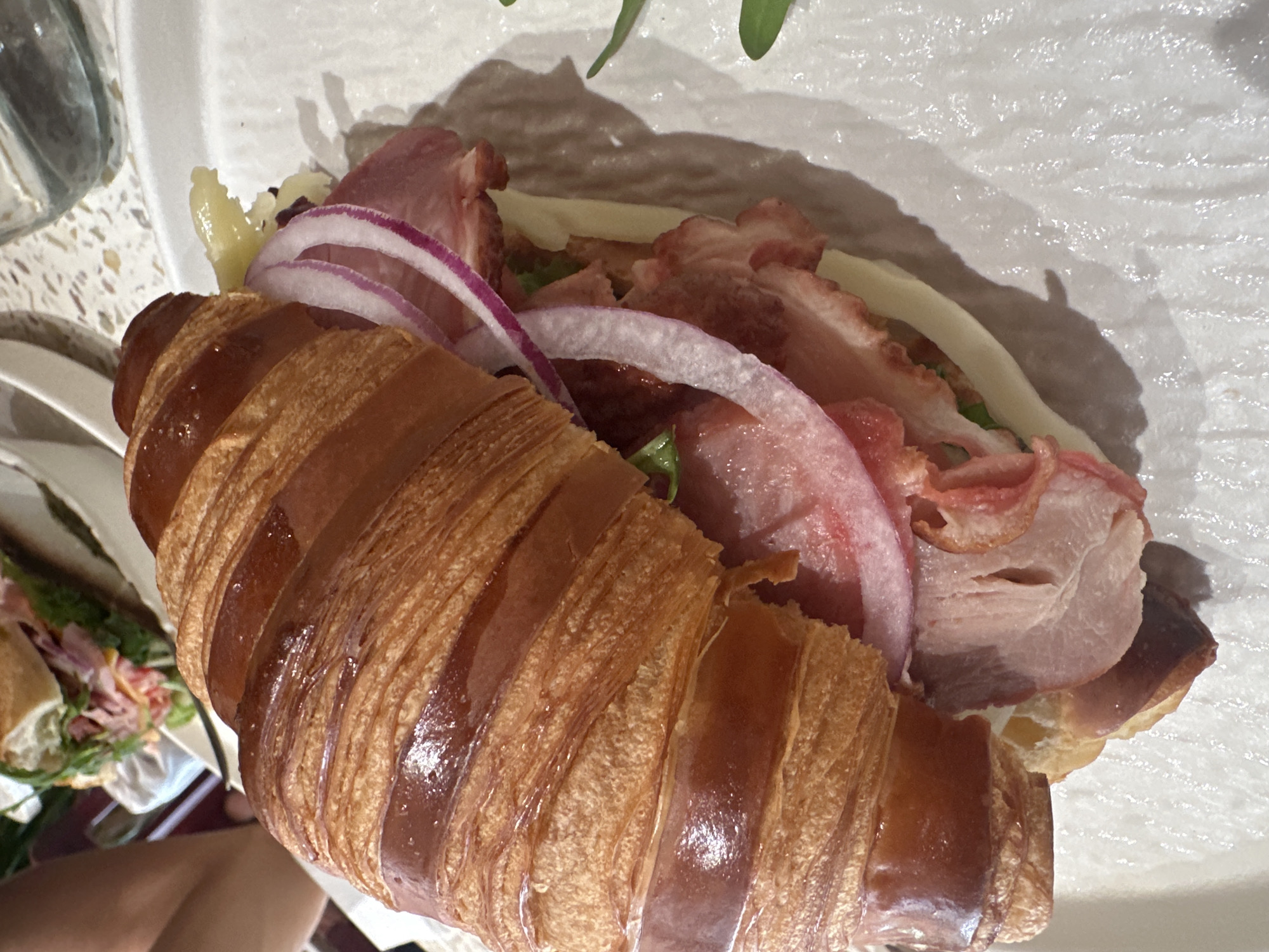 A croissant sandwich filled with bacon, red onion, cheese, and lettuce rests on a white plate. Part of another sandwich is visible in the background. The location is inferred to be in Bac Nha Trang Ward, Vietnam, but this is not explicitly confirmed by the image.