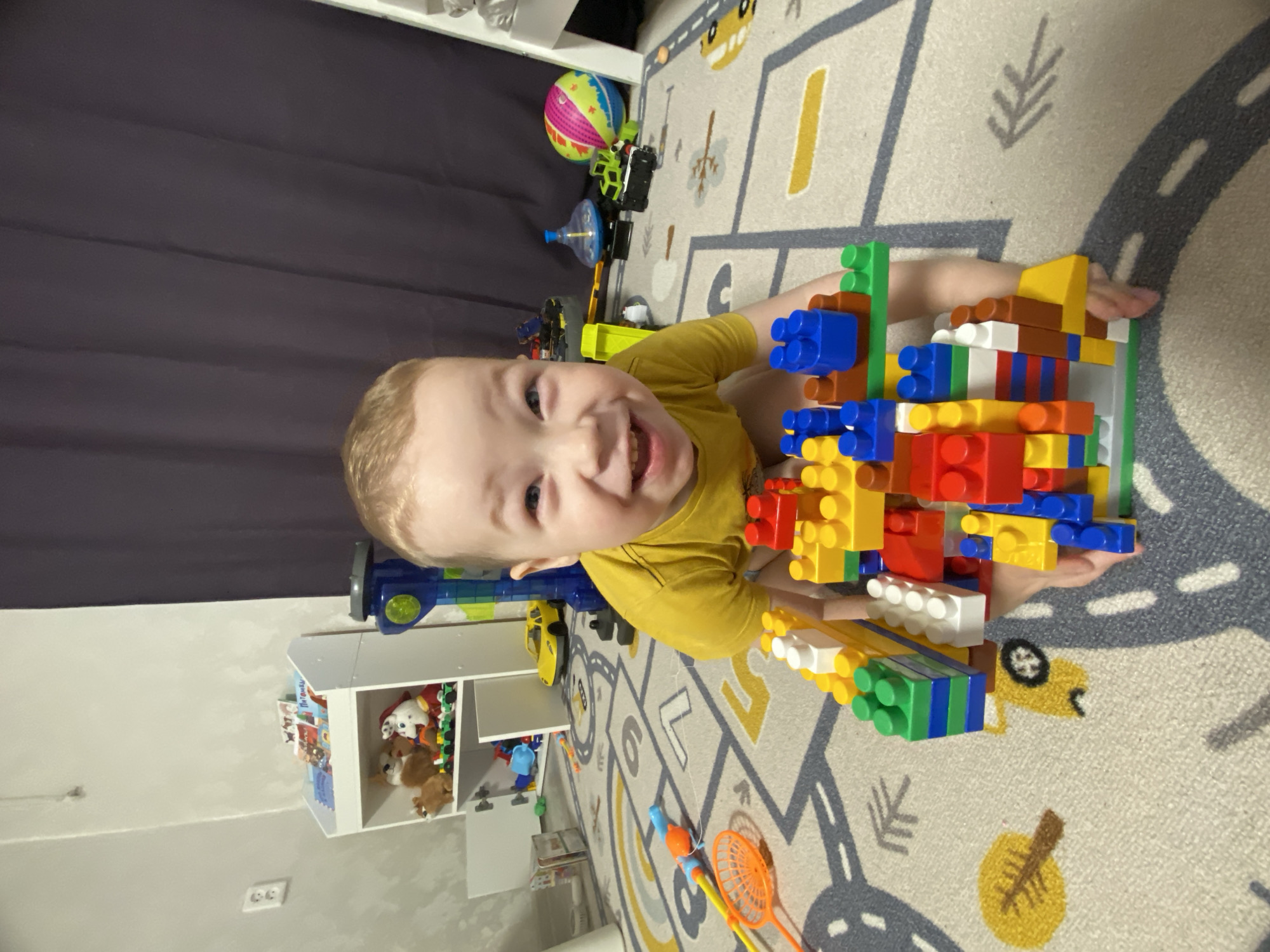 This media file captures a young child playing cheerfully indoors in Sochi, Russia. The child, with light hair and wearing a yellow t-shirt, is positioned on a light-colored rug adorned with a playful road and vehicle pattern. They are looking directly at the camera with a wide, open-mouthed smile, exuding joy and engagement.
The main activity involves the child playing with large, colorful building blocks. They are holding a substantial, multi-colored structure made from red, blue, yellow, green, white, and brown blocks. The child's hands are visible, supporting their creation.
The setting appears to be a child's room or play area. In the background, a dark grey fabric, possibly a curtain or wall covering, is visible. To the left, a white shelving unit contains various toys and books, including stuffed animals like a white dog and other brown plush toys. One of the books displays clear Cyrillic text, "Потешки," which translates to "nursery rhymes" or "lullabies." Other toys are scattered around the rug, such as a vibrant striped ball, a green and black toy vehicle, a blue spinning top, a yellow toy car, and orange rackets with a toy fishing rod. The room is brightly lit, indicative of daytime or well-lit artificial interior lighting. The overall scene conveys a happy, playful, and comfortable environment for the child.