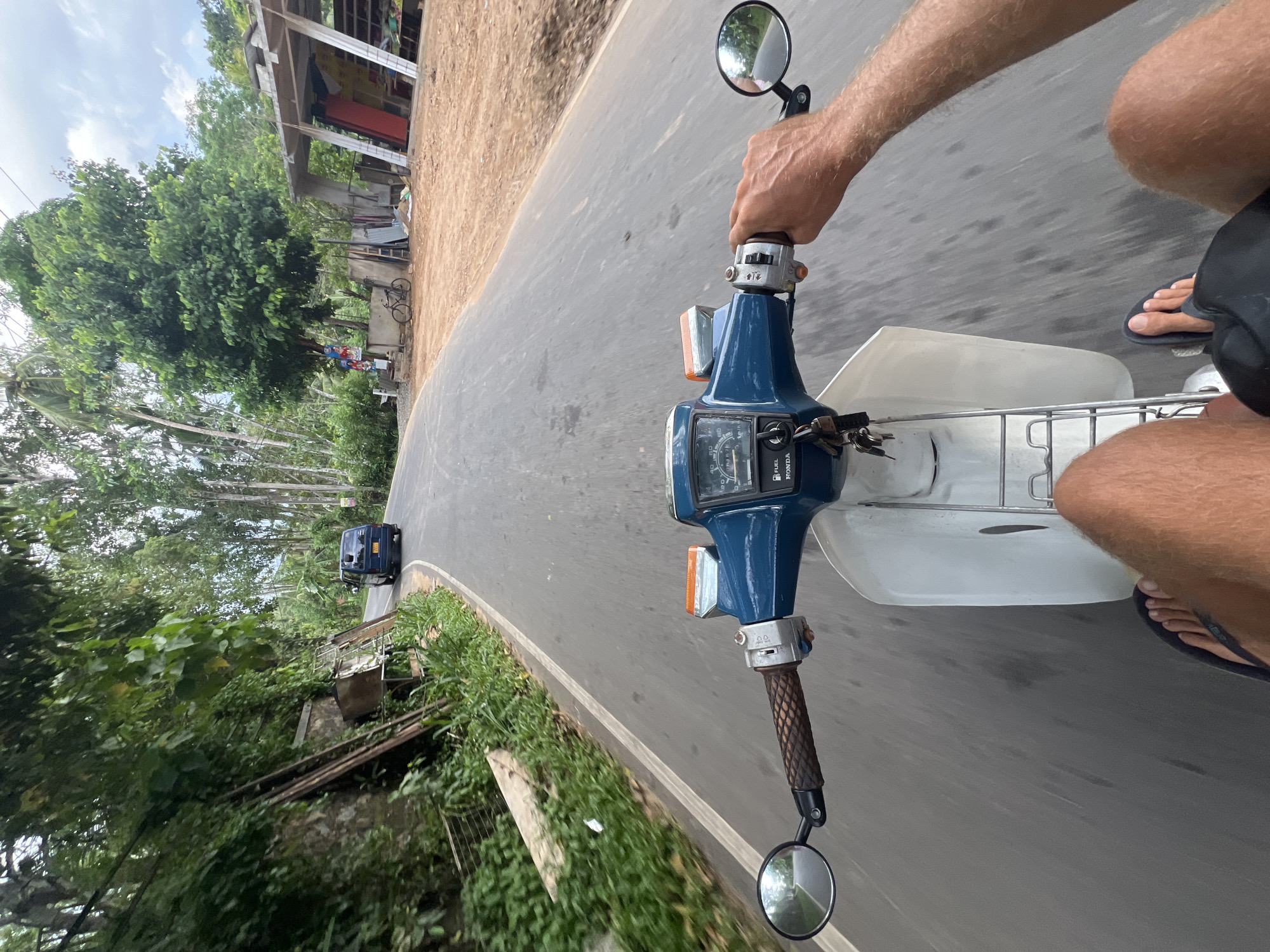 The image, taken from the first-person perspective of a person riding a blue Honda motorcycle, shows a road in Angulugaha Junction, Sri Lanka. The rider's hand is on the right handlebar. The road curves to the left, and a blue vehicle is visible in the distance. Buildings and vegetation line the roadside. The sky is overcast.