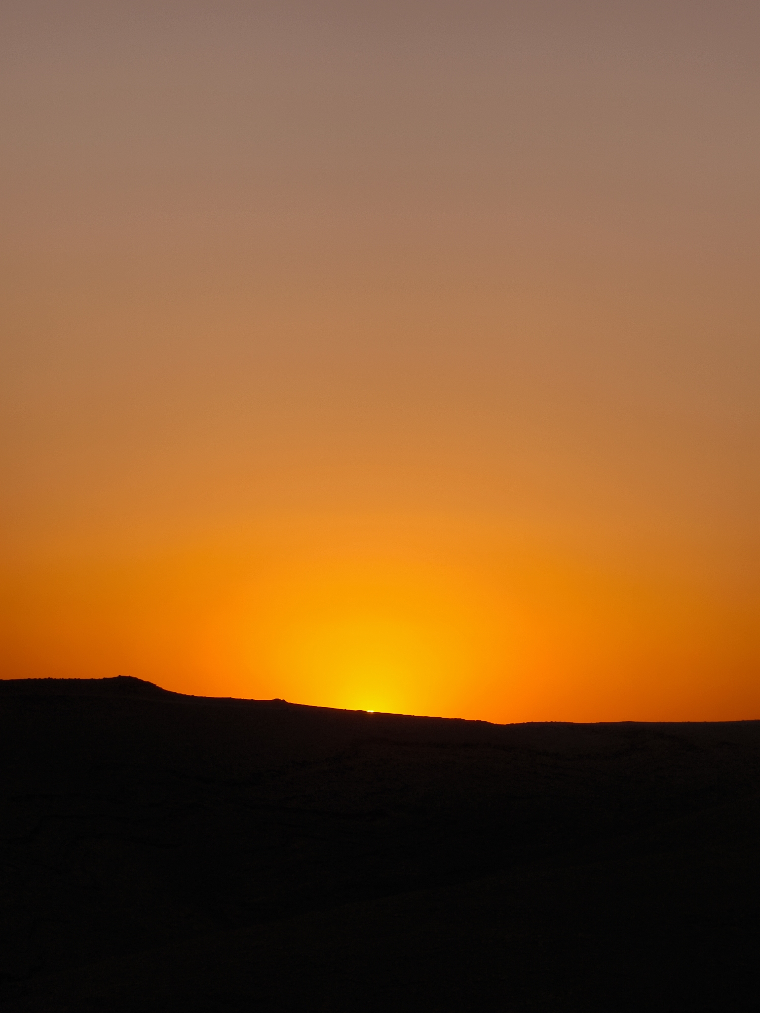 This is a photograph of a sunset in Keidar, Palestinian Territories. The scene is dominated by the sky, which transitions from a soft, light peach color at the top to a vibrant, fiery orange near the horizon. The sun is partially visible, a bright, glowing orb peeking over a dark, silhouetted landscape. This landscape appears to be a rolling hill or mountainous terrain, rendered in deep black due to the backlight of the setting sun. There are no visible people, objects, or specific structures within the frame. The focus of the image is the natural phenomenon of the sunset and its dramatic interplay of light and shadow against the arid terrain.