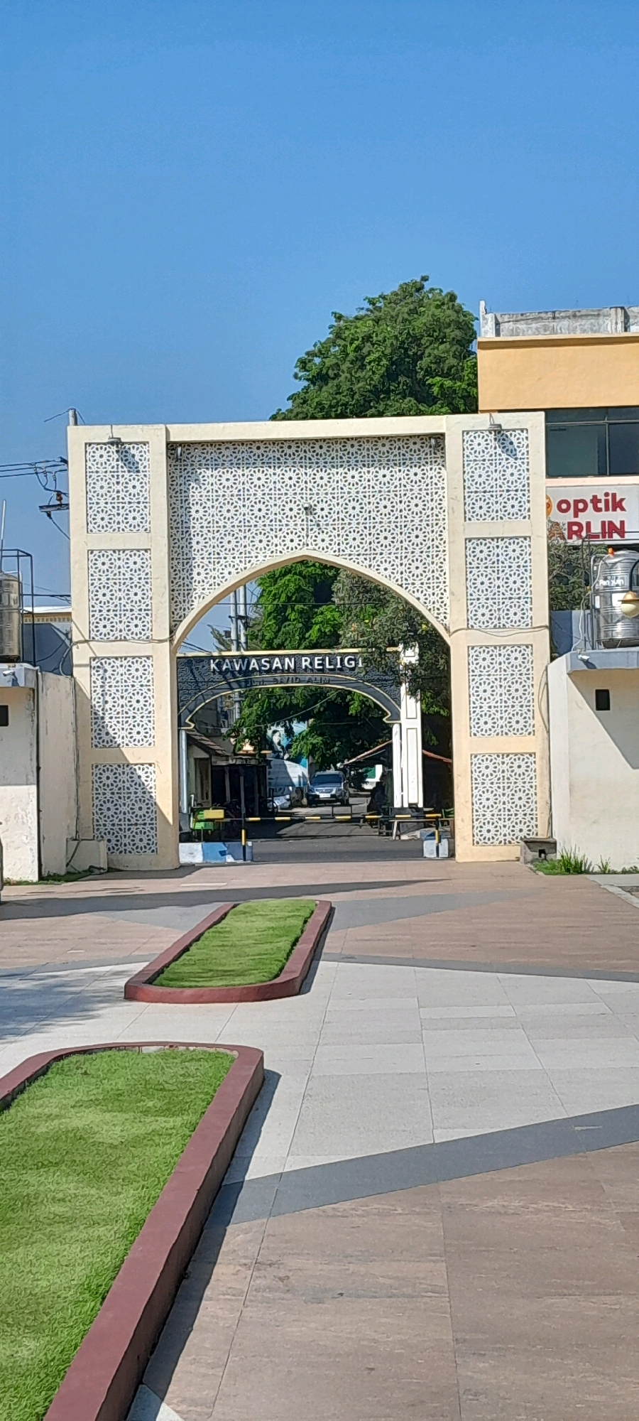 This is a full vertical shot of an arched gateway to a religious area in Pasuruan, Indonesia. The gateway is made of concrete with decorative Islamic geometric patterns on the side pillars and above the arch. The words "KAWASAN RELIGI" are visible on a dark blue banner under the arch. Beyond the arch, a street is visible with trees, buildings, cars, and barriers. In the foreground, there are two rectangular planters with manicured green grass. The ground is paved with large tiles. The sky is clear and blue.