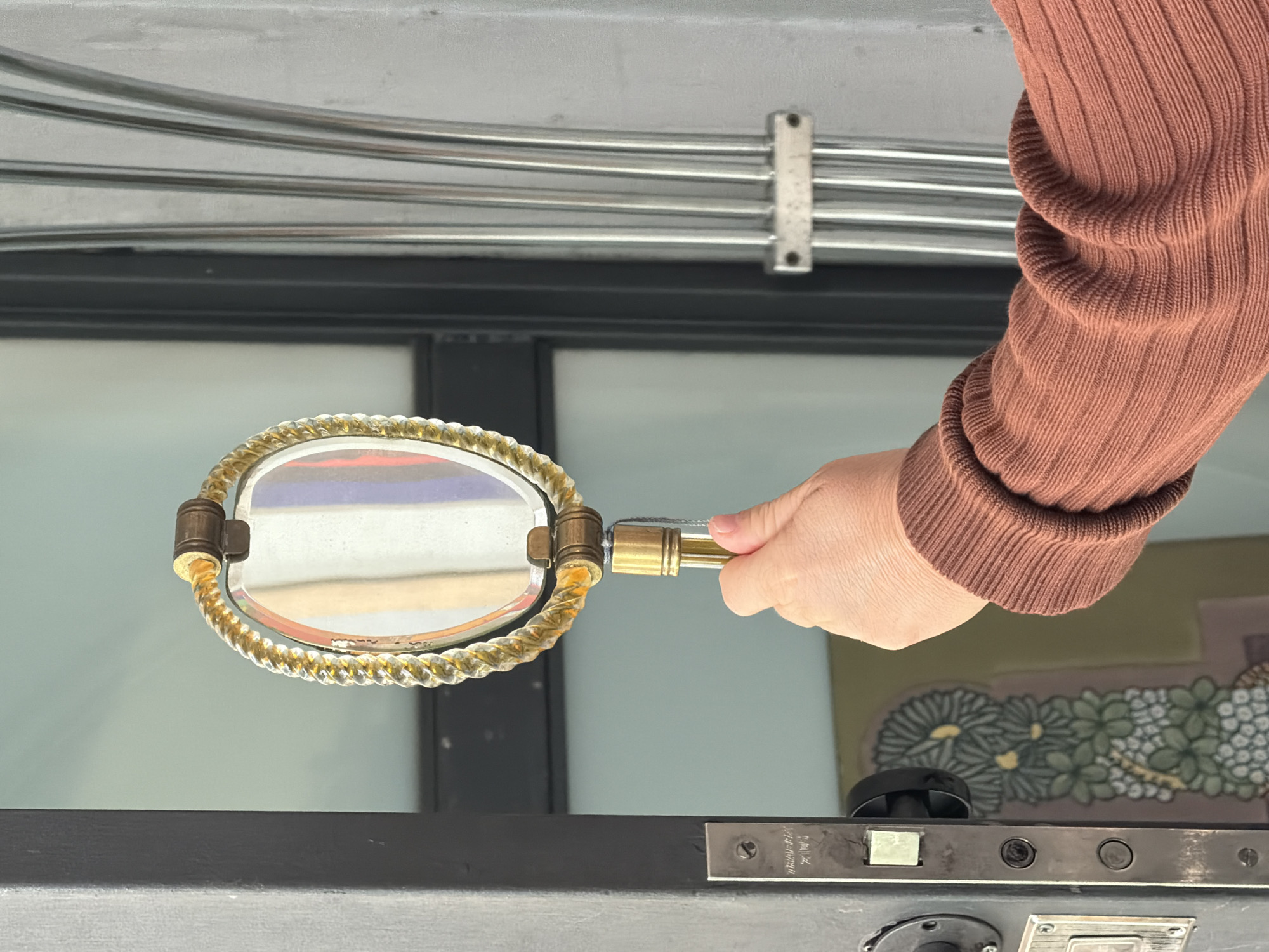 A hand holds a small, ornate mirror up to a dark-colored door, likely for inspection. The hand is attached to an arm wearing a brown, ribbed sweater. The door has a standard lock mechanism with visible screws. Behind the door, through the mirror's reflection, the sky and a building are visible. Above the door and hand, there are metal pipes secured with a white bracket. A mural is partially visible behind the door, featuring flower-like patterns in shades of green, white, and purple. Given the architectural style and mural designs, the location may be a building in Buenos Aires, Argentina.