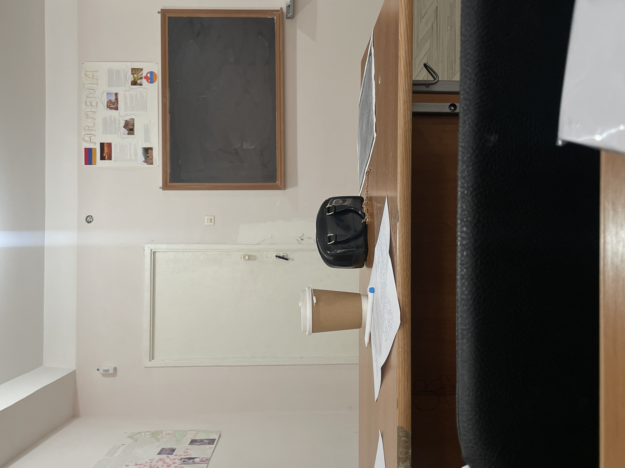 The image shows a room in Yerevan, Armenia, indicated by the "Armenia" poster on the wall. The room features a chalkboard, a door, and a desk. On the desk are a coffee cup, a black purse with a gold chain, and papers. A black office chair is partially visible on the right. The room has light pink walls.