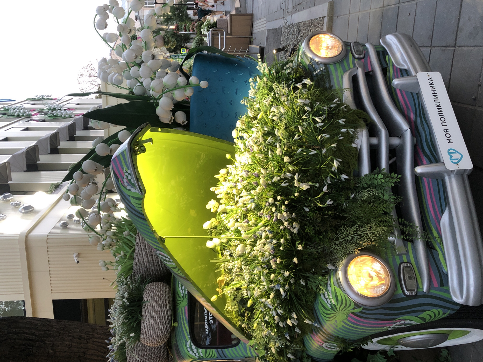 This image captures a creatively decorated vintage car, possibly a Volkswagen Beetle, adorned with an abundance of white flowers, resembling lily of the valley, and lush green foliage. The car itself is painted with a vibrant, swirling green and blue pattern accented with pink stripes, giving it a whimsical appearance. The hood is open, revealing a bright lime green interior.
The car is situated outdoors, on what appears to be a paved area, likely a sidewalk or pedestrian walkway. In the background, there's a modern building with clean lines and large windows, suggesting an urban setting. A hint of foliage and trees further contextualizes it as an outdoor scene. The lighting suggests it might be daytime, with the sun casting a warm glow on the flowers and the car's headlights, which are illuminated.
Attached to the front bumper is a white sign with a blue logo and text in Cyrillic, which reads "МОЯ ПОЛИКЛИНИКА" (My Clinic). Another piece of visible text, partially obscured on the side of the car, appears to be in Cyrillic as well, possibly part of a slogan.
The overall impression is one of a festive or decorative display, perhaps for an event or a business promotion, given the signage. The abundance of flowers and the cheerful colors evoke a sense of springtime or celebration. There are no people visible in the frame, focusing the attention entirely on the decorated vehicle and its surroundings. The image is oriented vertically, suggesting it was taken with a mobile device or oriented for a portrait display.