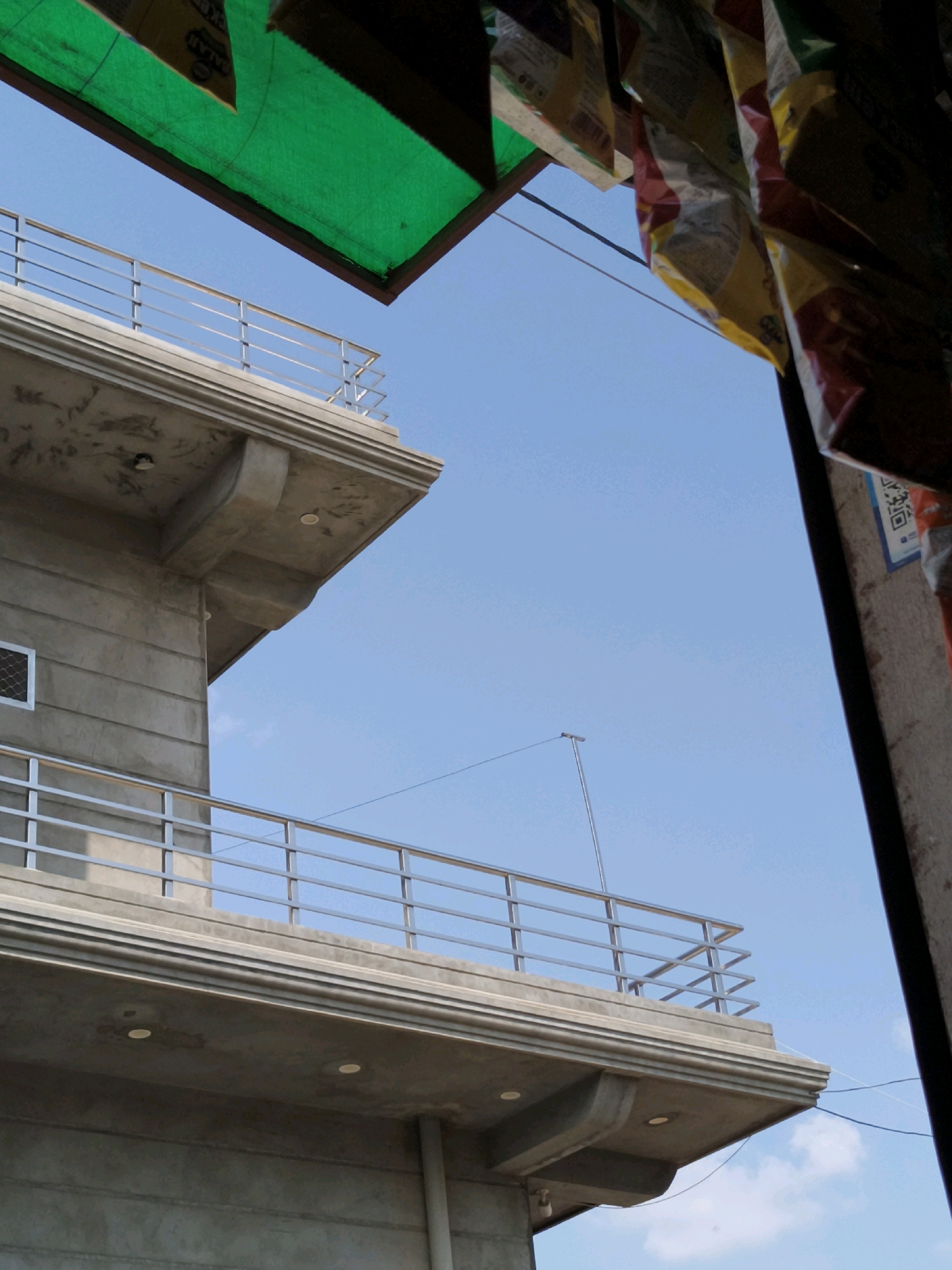 The image shows a close-up, low-angle view of the upper levels of a concrete building with a modern design, against a clear blue sky. The building features multiple balconies with horizontal metal railings. The concrete surfaces appear unfinished, revealing the texture of the material. Two small, round light fixtures are visible on the underside of one of the balconies. A single window with a metal mesh is present on the side of the building.
In the foreground, obscuring the top portion of the sky and the building, are elements that suggest a street-level market or vendor stall. A green tarp-like material is visible at the top left, and what appear to be hanging bags or packaging, possibly for sale, are seen on the right. The packaging has various colors, including yellow, red, and white, and some contain text and graphics, including what looks like a QR code. Thin wires are also visible spanning across the scene in the sky.
The scene is set in Taranagar, India. The overall impression is of an urban environment, likely a commercial area, with ongoing construction or renovation evident in the building's appearance. No people are visible in the image.