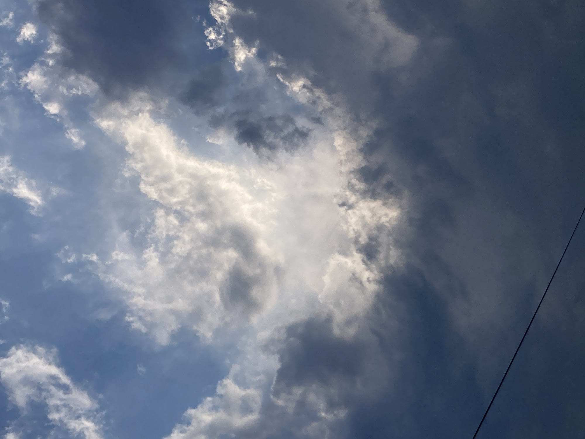 The image captures a sky filled with dramatic clouds above Kemer, Turkey. A central area of bright, illuminated clouds forms a roughly oval shape, suggesting sunlight breaking through or reflecting off the cloud formations. Surrounding this bright area are darker, more ominous clouds, creating a stark contrast. A thin, dark line, likely a power line or cable, bisects the lower portion of the image diagonally from left to right. The overall impression is of a dynamic sky with varying light conditions, possibly indicating an approaching storm or the aftermath of one.