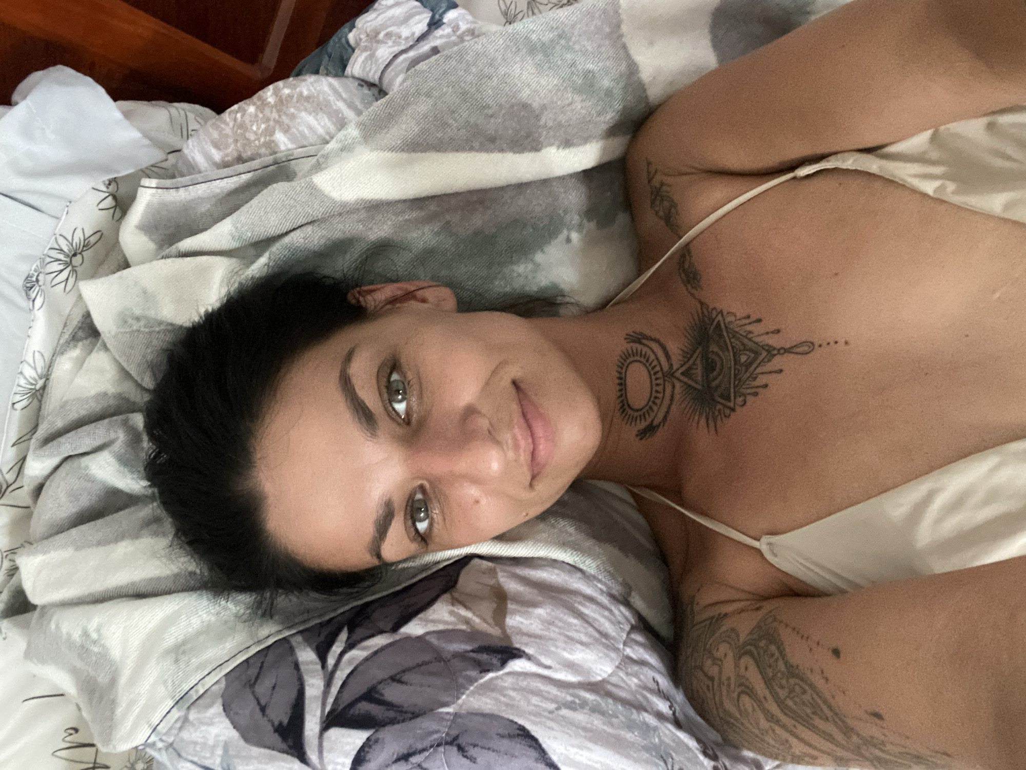A woman with dark hair and tattoos lies on a bed in Mũi Né, Vietnam. She is wearing a white bikini top and looking at the camera with a slight smile. The bed has a patterned blanket. The room appears to be a bedroom.