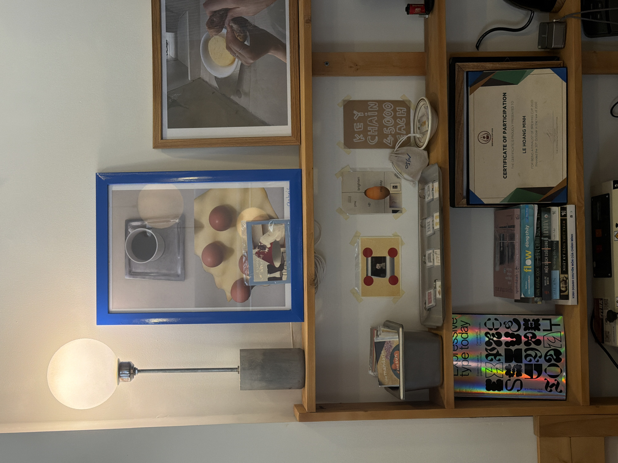 The image presents a curated shelf display, with a light to the lower left. At the top left is a framed photo of hands preparing food, next to a framed blue-bordered picture of eggs, a coffee cup and a photo. The shelf is divided into sections containing various small items and artworks, including a price tag for "Key Chain 4-5.000 Each". There are objects like a respirator mask, small photos, and containers of books. The lower shelf houses a book titled "Expressive Type Today". A Certificate of Participation awarded to "Le Hoang Minh" for brewing is also present. Several books, with titles in both English and Vietnamese, are stacked on the shelf. Đà Nẵng, Vietnam is implied by the writing on some of the books.