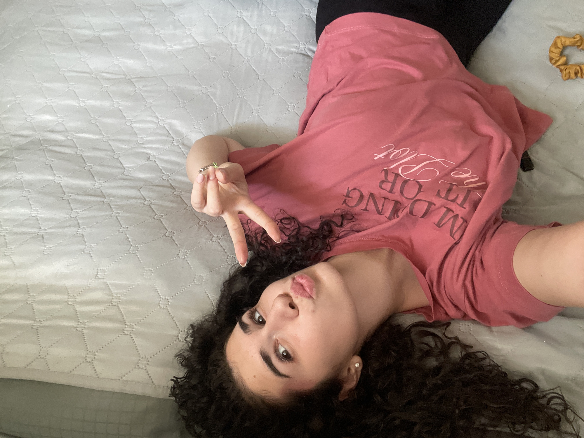 A young woman lies on her back on a light grey patterned bedspread, wearing black leggings and a dark pink t-shirt with white lettering. Her dark, curly hair fans out around her head. She is making a peace sign with her right hand and has a ring on her index finger. She is looking at the camera with her lips pursed. A scrunchie is visible in the upper right corner of the image. The location is not discernible.