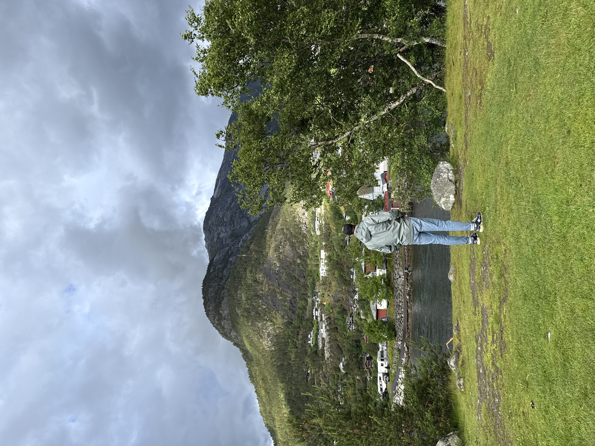 A man stands with his back to the camera, looking towards a scenic view in Eidfjord, Norway. He is wearing a light gray hooded jacket and blue jeans. The man is positioned on a grassy area in the foreground, with a body of water to his left and a hillside dotted with houses and trees behind it. The hillside is covered in greenery and rocky outcrops, and a large mountain rises in the background under a cloudy sky. A birch tree with green leaves is visible to the right of the man. Several white RVs and cars are parked along the edge of the water, suggesting a campsite or parking area. The overall atmosphere is tranquil and picturesque, capturing the natural beauty of the Norwegian landscape.