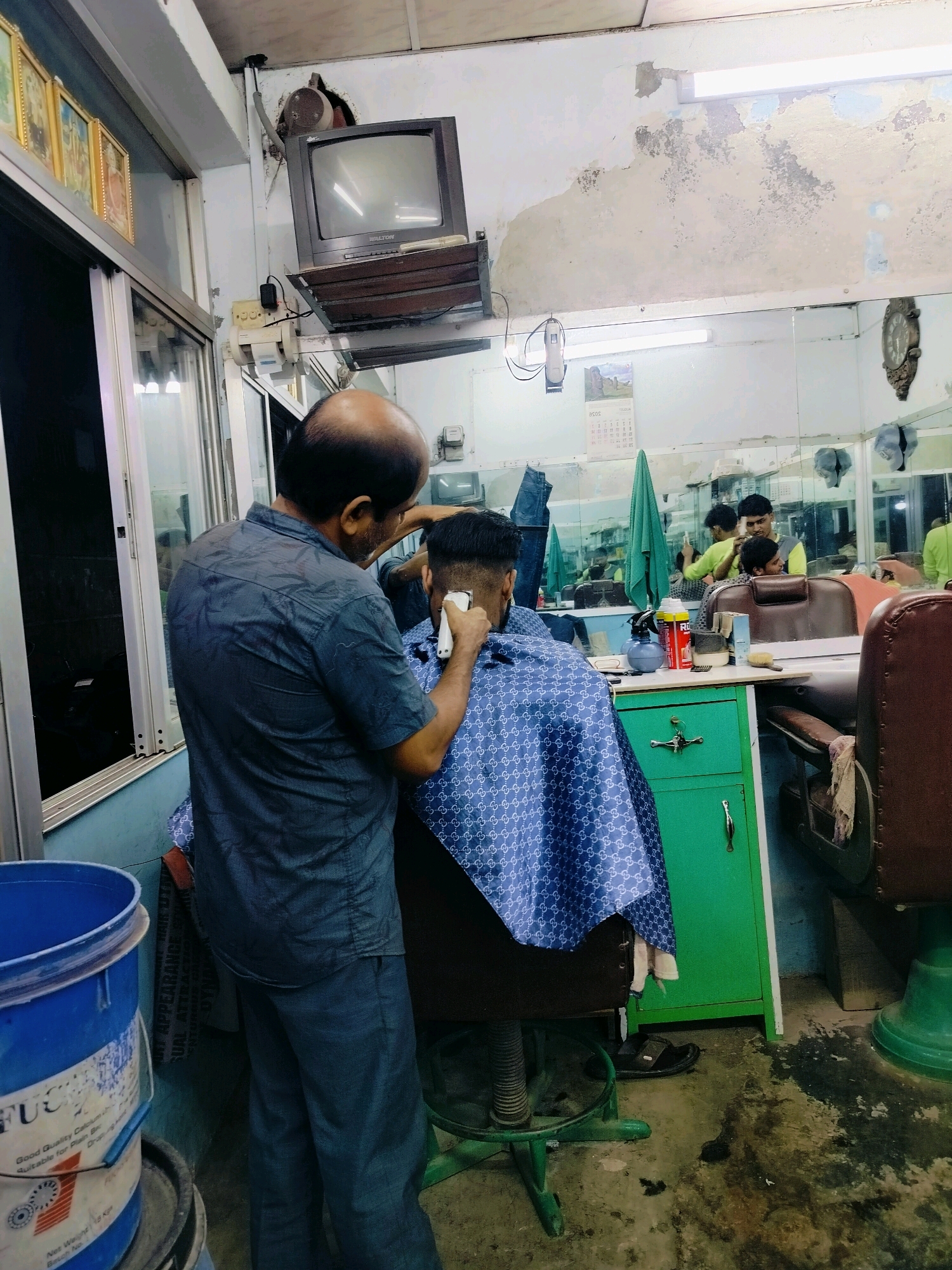 The image captures a scene inside a barber shop located in the bustling city of Dhaka, Bangladesh. In the foreground, a bald man, presumably the barber, is focused on giving a haircut to a young man seated in a barber chair. The barber is wearing a dark blue, patterned short-sleeved shirt and dark pants. He is holding an electric hair clipper in his right hand, carefully trimming the hair on the back of the customer's head. The customer is wearing a blue cape patterned with white interlocking G logos, typical of Gucci.
The barber shop appears to be modestly equipped, with mirrors lining the walls reflecting other parts of the shop and its occupants. A television set is mounted on the wall in the upper left corner, and a clock hangs on the right wall. A green cabinet with silver handles is positioned behind the barber's chair. Various barbering tools and supplies, such as a spray bottle, cans, and brushes, are visible on a counter. In the lower left corner, a blue plastic bucket with some labeling is present. The floor is concrete, showing signs of wear and some scattered debris. The overall lighting suggests an indoor setting, likely illuminated by overhead fluorescent lights. The ambient environment hints at a typical, everyday scene in a local Dhaka establishment.