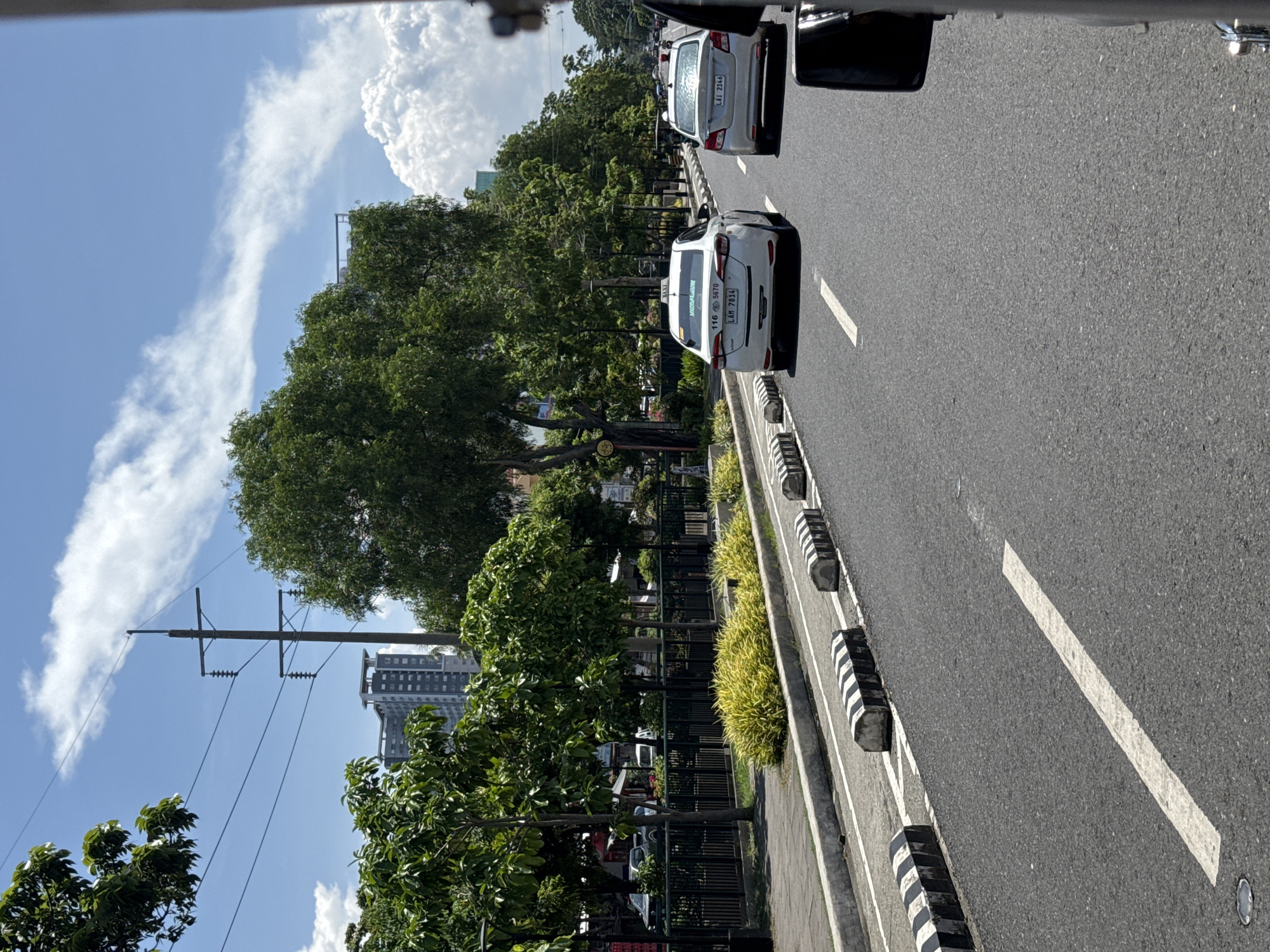 The image captures a sunny daytime scene on a multi-lane road in Davao City, Philippines, viewed from inside a vehicle. The perspective looks forward, with the road stretching into the distance.
In the foreground and midground, two vehicles are visible in a lane. The closest car is a white sedan taxi, clearly displaying "TAXI" on its rear, along with what appears to be a taxi number "116 5570" and the word "MISSFLAVOR" on its back window. Its license plate reads "LAM 7814." Directly behind it is a silver sedan with the license plate "LAI 2146." Both cars appear to be either stopped or moving slowly, suggesting traffic or a stop.
To the left of the road is a median strip, delineated by black and white striped concrete barriers and a black metal fence. This median is extensively planted with lush green trees and vibrant yellow-green shrubs. Beyond the trees, several multi-story buildings are visible in the background, hinting at an urban environment.
The sky above is bright blue with scattered white cumulus clouds, indicating clear and sunny weather, likely around midday or early afternoon given the strong illumination and shadows. A wooden utility pole with multiple cross arms and electrical wires stands prominently against the sky on the left side of the frame. The asphalt road surface features a clear white lane marking on the right side. The overall scene depicts a typical busy street in the city.