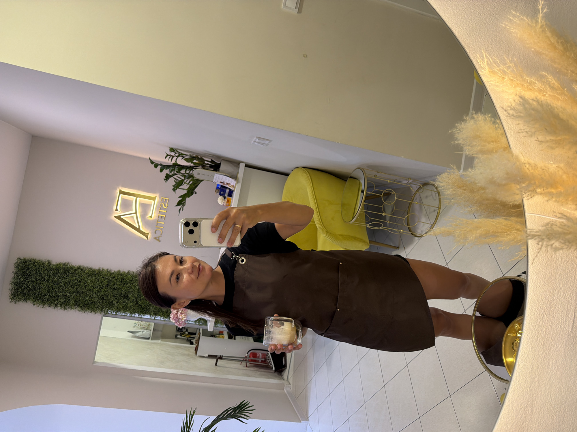A woman in a brown apron and black shirt is standing in what appears to be a salon or spa, holding a clear glass with a frothy beverage. She is taking a mirror selfie with a white smartphone. In the background, a large mirror reflects the room, showing a vertical green plant installation on the wall, a yellow chair, and a gold-colored accent table. Above the plant, a glowing neon sign with the letters "EA ESTETICA" is visible. The floor is tiled with white square tiles. Another reflection in the mirror shows a doorway and a fire extinguisher. In the foreground of the mirror's reflection, decorative dried pampas grass is arranged. The lighting is bright and warm, suggesting an indoor environment. The overall atmosphere is clean and modern.