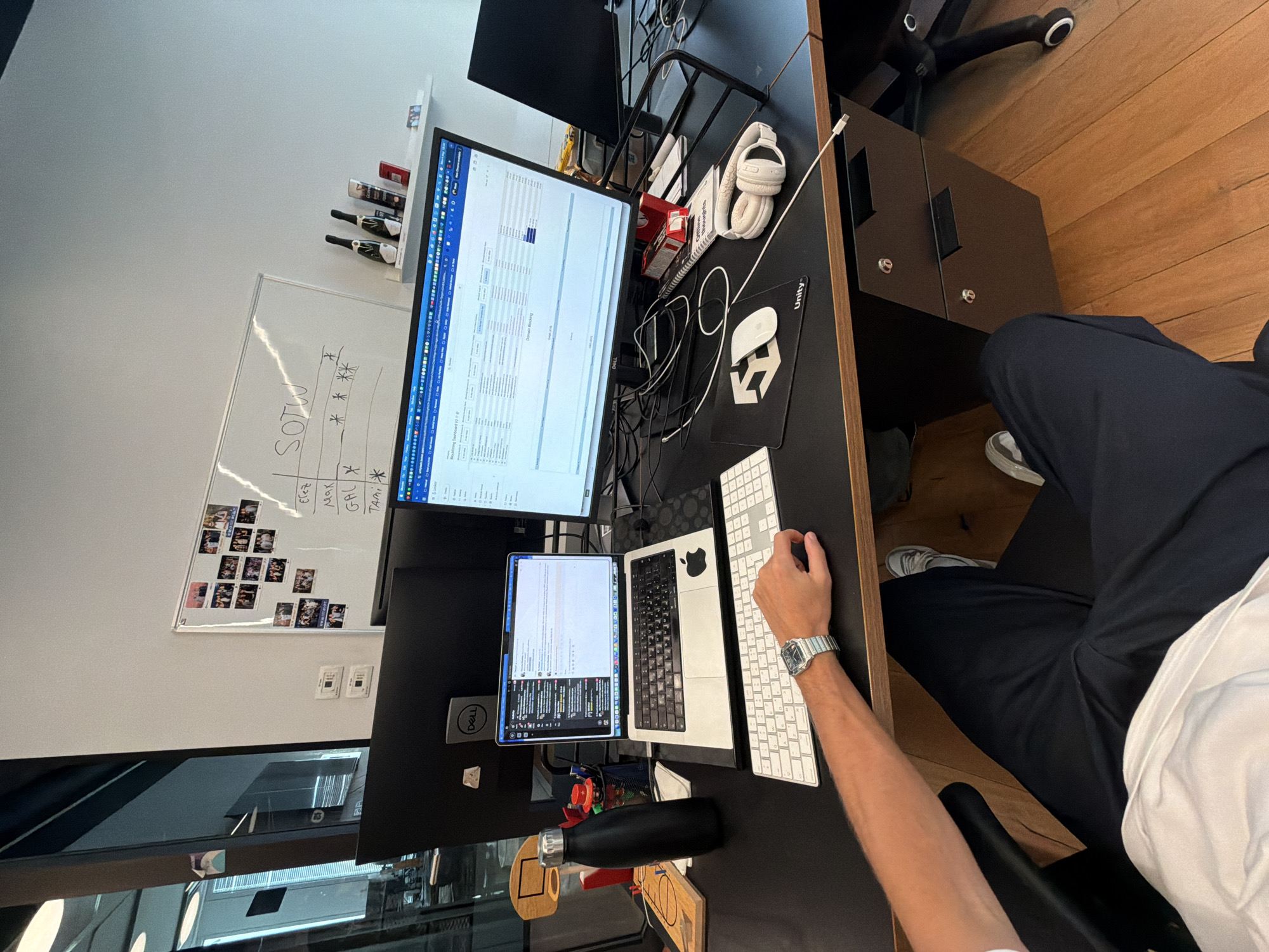 The image depicts a workspace, likely an office, with a desk cluttered with technology and personal items. A person is seated at the desk, using a white keyboard and wearing a silver wristwatch. On the desk, there are two computer monitors, a laptop, a mousepad with a "Unity" logo, and headphones. There's also a shelf with bottles and a whiteboard with writing and photographs. The floor appears to be made of wood. The location is likely within an office building in Sdot Negev Regional Council, Israel.