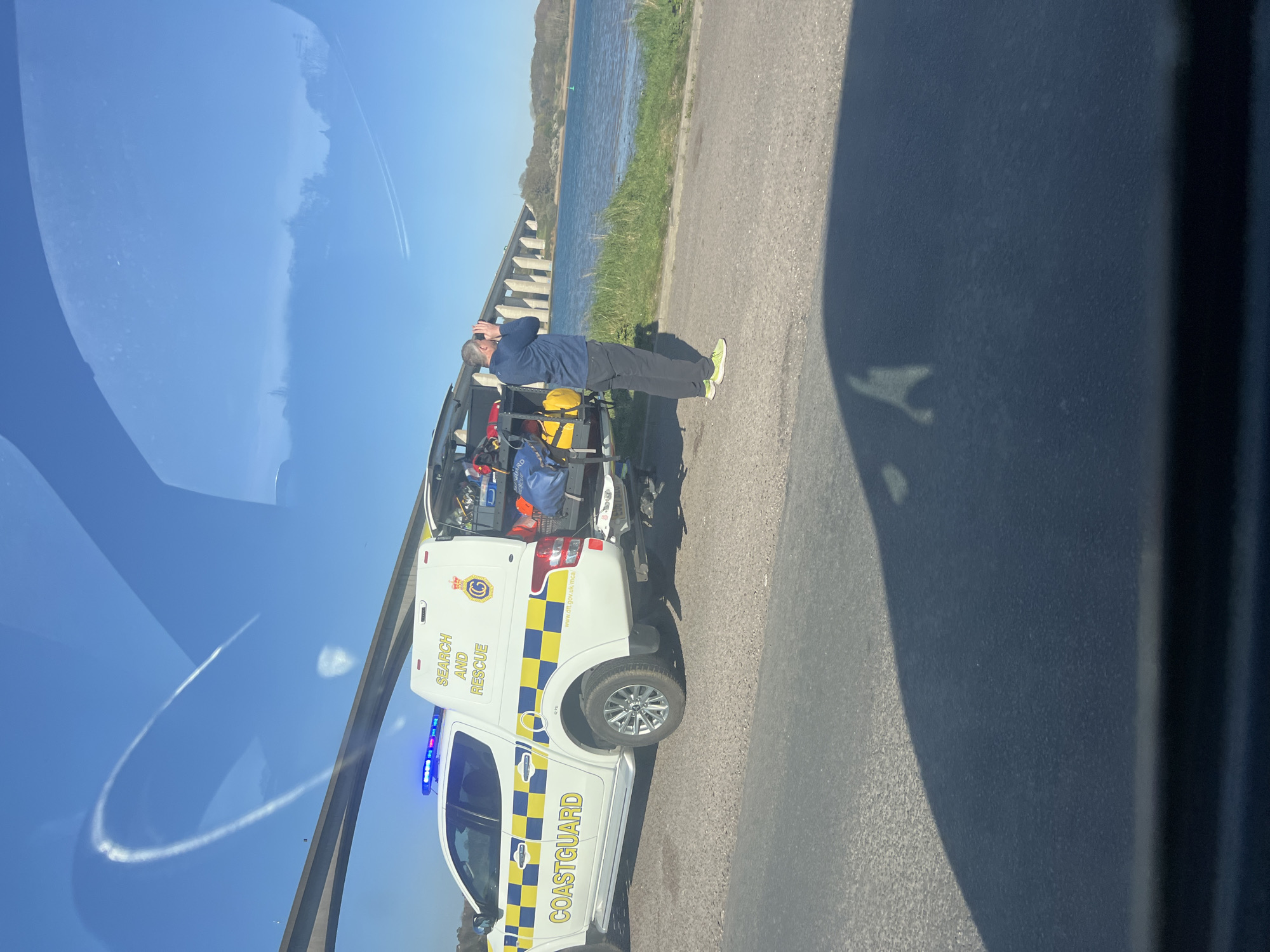A Coastguard search and rescue vehicle is parked on a road adjacent to a body of water. A man stands at the rear of the vehicle, holding binoculars to his eyes. The vehicle is white with blue and yellow checkered markings and the words "COASTGUARD" and "SEARCH AND RESCUE" are clearly visible. The back of the vehicle is open, revealing equipment inside. The scene is outdoors, with a clear sky and a body of water. The location is likely Babergh, United Kingdom, given the Coastguard marking.