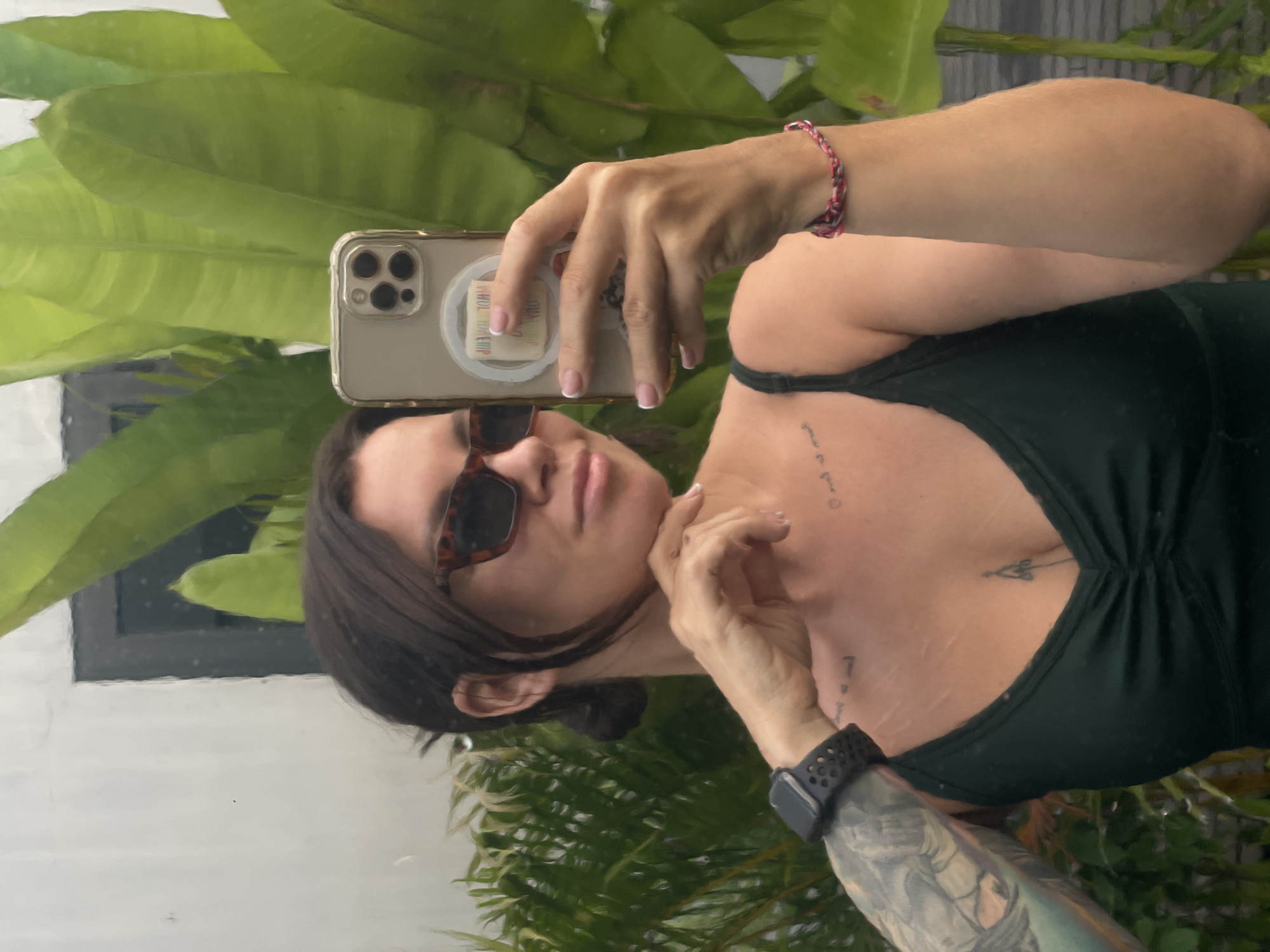 In Kediri, Indonesia, a woman with dark hair pulled back is taking a selfie in a mirror. She is wearing sunglasses and a dark green tank top. Her left arm is visible, showing a tattoo and a black smartwatch. A pink and red braided bracelet adorns her right wrist. She has a small tattoo on her chest and other faint markings on her upper chest. The background consists of lush green foliage and a light-colored wall with a dark window frame. The reflection also reveals a smartphone in her hand, held up to capture the image.