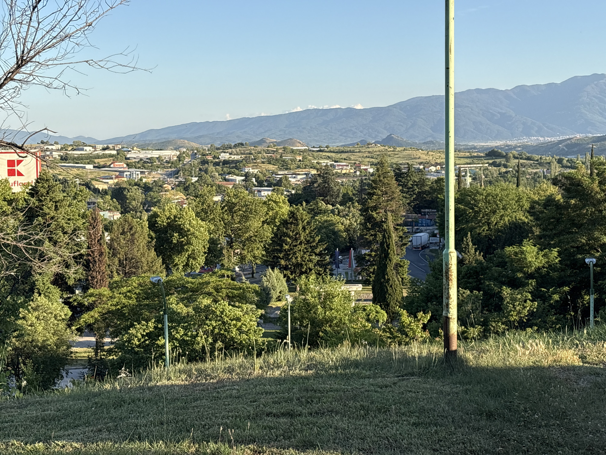The image presents a landscape view from a slightly elevated grassy area, possibly a hillside or park, overlooking a cityscape and mountain range. The foreground shows green grass with some weeds and vegetation. A green metal pole is located to the right of the image frame.
In the middle ground, there is a dense collection of trees and greenery, partially obscuring the view of the city. Interspersed among the trees are other lampposts.
In the distance, the cityscape consists of buildings of varying heights and architectural styles, typical of a residential or mixed-use area. Beyond the city, a mountain range stretches across the horizon under a clear, light blue sky. The city is Sandanski, Bulgaria.