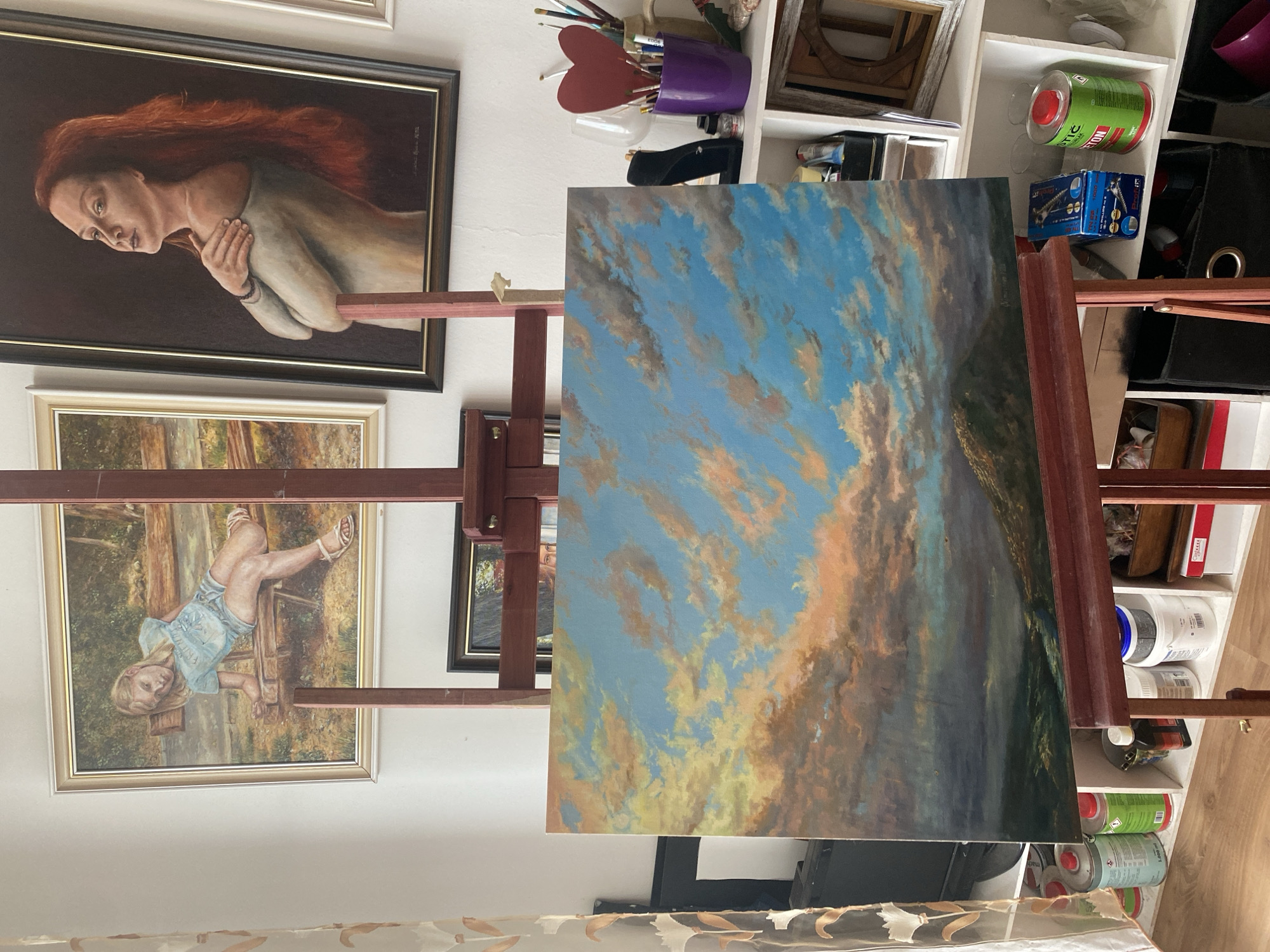 The image depicts an art studio, possibly in Bílina, Czechia, filled with paintings. In the foreground, a landscape painting of a sky with dramatic clouds over rolling hills is displayed on a wooden easel. To the left of the easel, a curtain with a floral pattern is visible. Behind the foreground painting, two framed portraits hang on the wall: one of a young girl sitting on a bench, and another of a woman with long red hair. A shelf unit to the right of the easel holds various art supplies, including paintbrushes in a purple container, cans of paint, and empty containers. The studio appears to be a personal workspace, with an assortment of items suggesting active artistic creation.