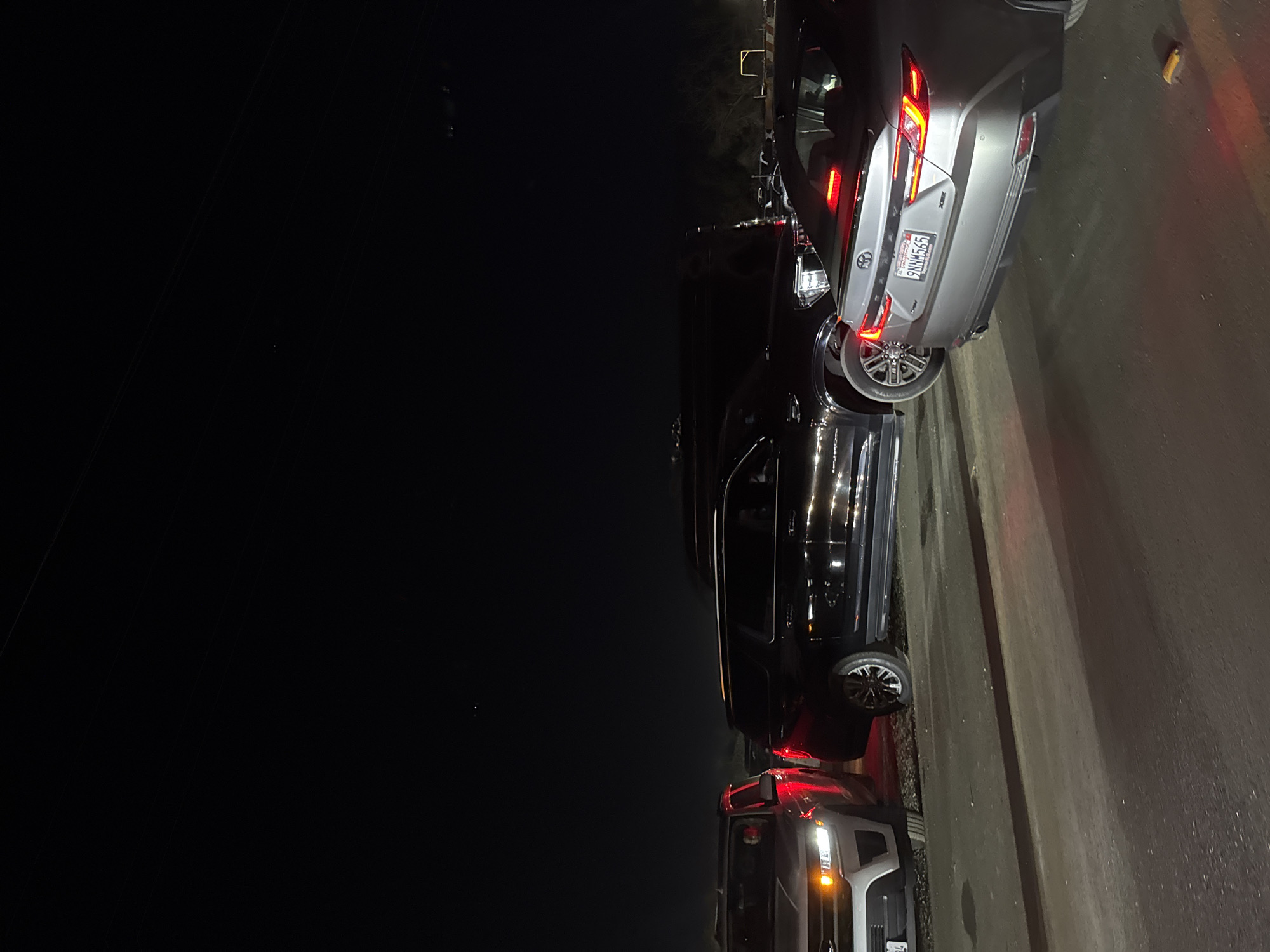 The image shows a night scene in Indio, California, United States of America. Several vehicles are parked alongside a road. From top to bottom, the first car is a gray sedan. A license plate that reads "9HNM565" can be seen on the vehicle. Underneath the sedan is a black SUV that is elongated in length. Below the SUV, a pickup truck can be seen, with the front of the car facing the road. The image is taken at night, and the background is obscured by darkness. The road appears to be made of asphalt.
