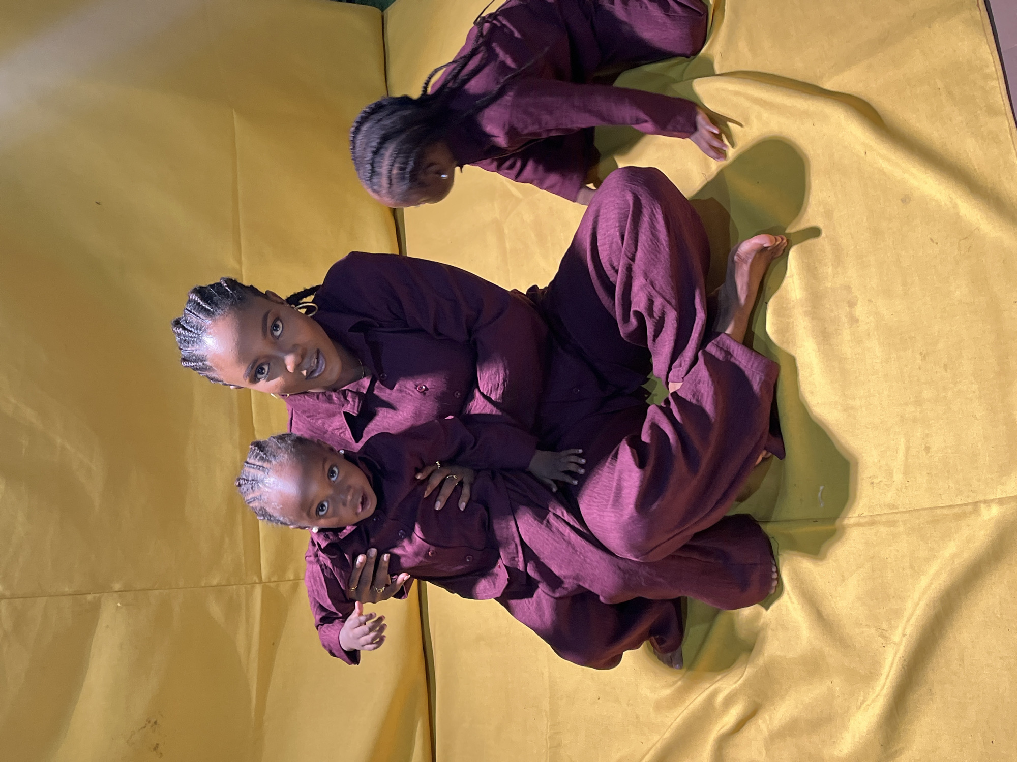 In a studio setting, possibly in Jos, Nigeria, a woman with braided hair sits on a yellow, creased backdrop. She is wearing a plum-colored matching set of a collared shirt and wide-leg pants. She holds a young child, also in a plum-colored matching set, on her lap. This child has braids and wears small earrings. Another young child, dressed similarly, is crouched to the right of the woman, facing away from the camera. The scene is lit, casting soft shadows on the yellow surface.