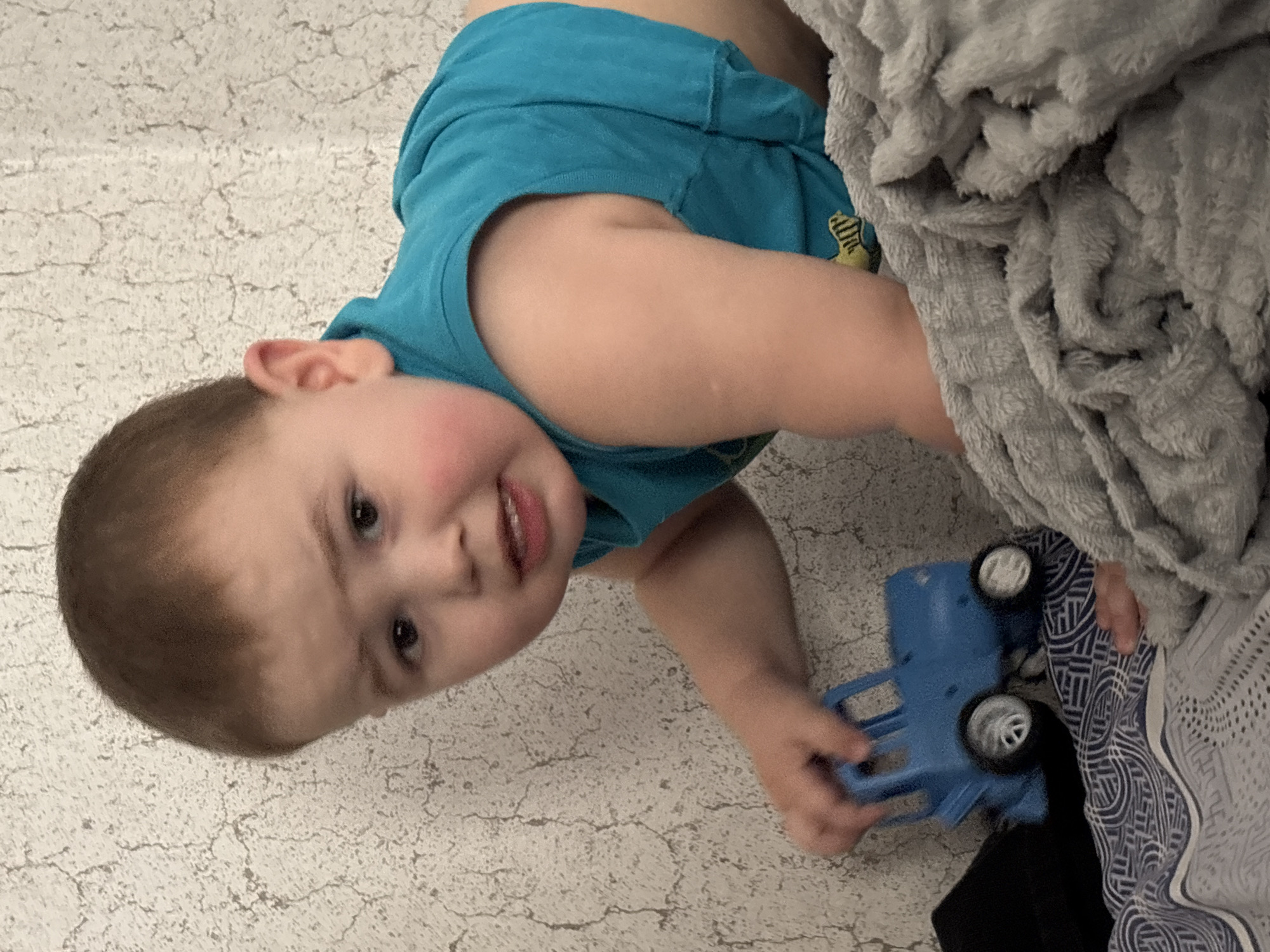 A toddler wearing a blue sleeveless shirt is lying on a cracked, off-white surface. The toddler is holding a blue toy truck. A gray, textured blanket is visible to the right of the child. The location is unknown, not necessarily Фоки, Russia.