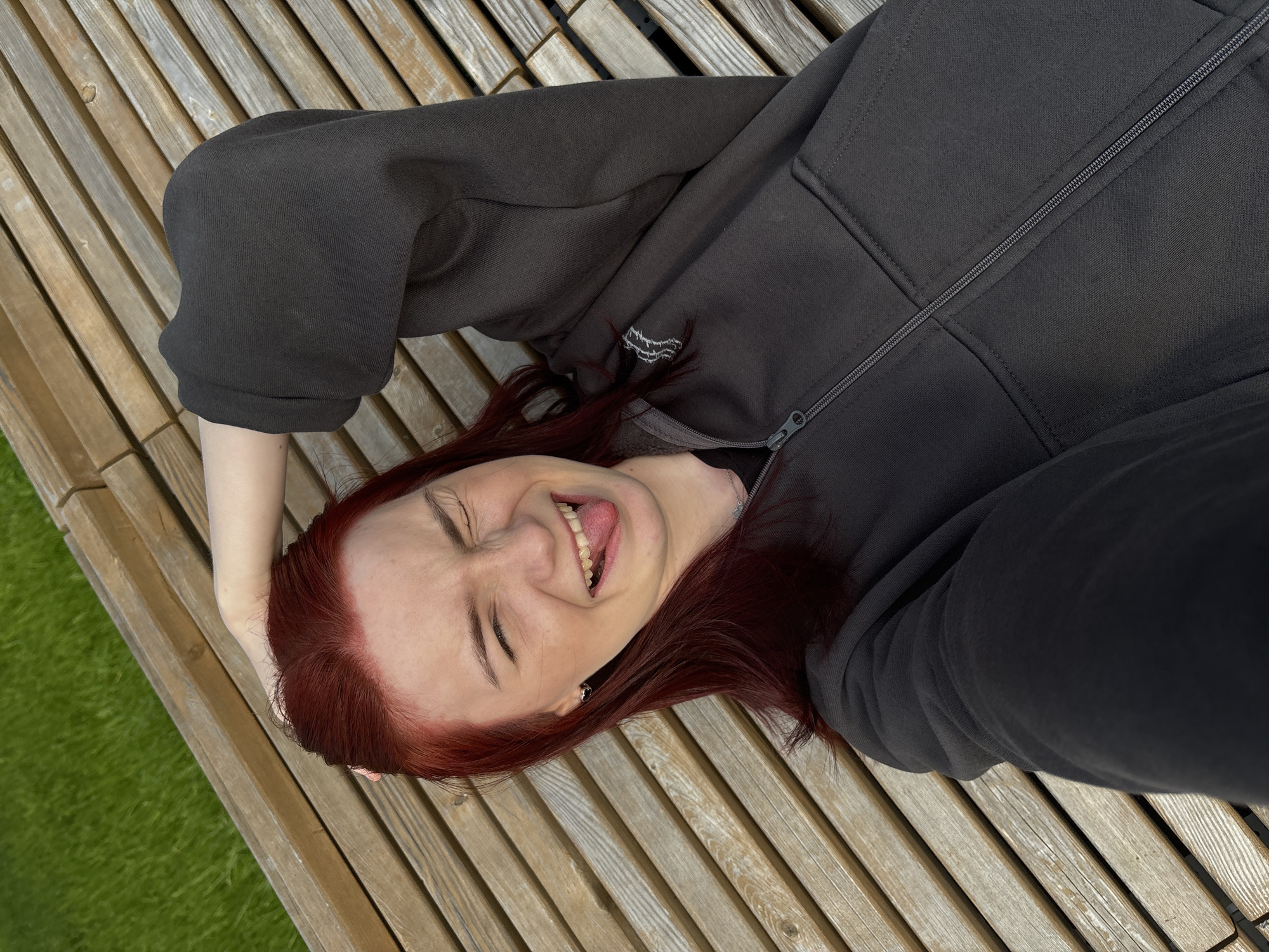 A young Caucasian woman with bright red hair lies on a wooden slatted surface with a bright green grassy area visible in the upper left corner. She is wearing a dark gray zip-up hoodie and has her right arm raised behind her head, with her hand resting on her hair. Her eyes are closed, her mouth is open in a wide smile, and her tongue is sticking out to the right. She is looking directly at the camera, suggesting she is taking a selfie. The wooden surface appears to be a bench or deck made of light-colored wood planks.