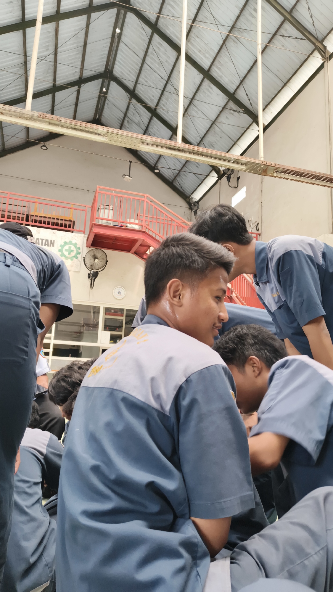 The image shows a group of young men wearing matching blue and gray work uniforms, likely in a workshop or industrial setting in Katrungan, Indonesia. The men appear to be gathered together, with some in the foreground looking towards a central point of focus, while others are bent over, possibly engaged in a task or discussion. The environment features a high, corrugated metal roof supported by a visible industrial framework and beams. Red metal railings and stairs are visible in the background, suggesting a multi-level structure within the building. A fan is mounted on the wall, and a sign with a green hazard symbol and text is also present. The lighting suggests an indoor space with diffused natural light, possibly from the roof or windows. The overall impression is one of a collective activity or training session within a functional industrial space.