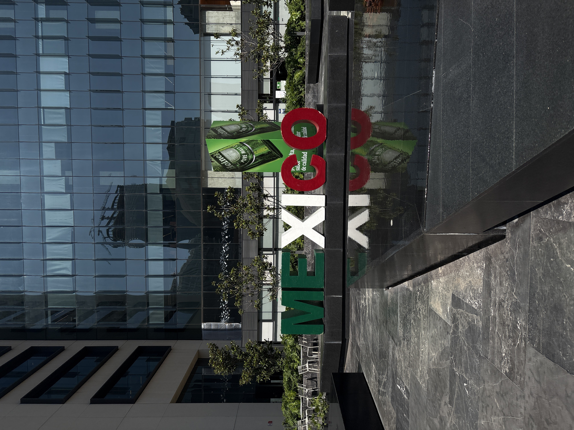 The image is an outdoor shot, possibly taken in Mexico City, Mexico, given the word "MEXICO" spelled out in large, colored letters. The “ME” is green, “XI” is white, and “CO” is red. The letters are positioned on a dark, patterned surface that appears to be a water feature, as reflections of the letters are visible on the surface. Behind the letters and water feature, there is a large Heineken advertisement. A modern, multi-story building with a glass facade is in the background, reflecting other buildings and structures. Small trees and landscaping are visible between the lettering and the building, and there are metal chairs and tables in a shaded area nearby.