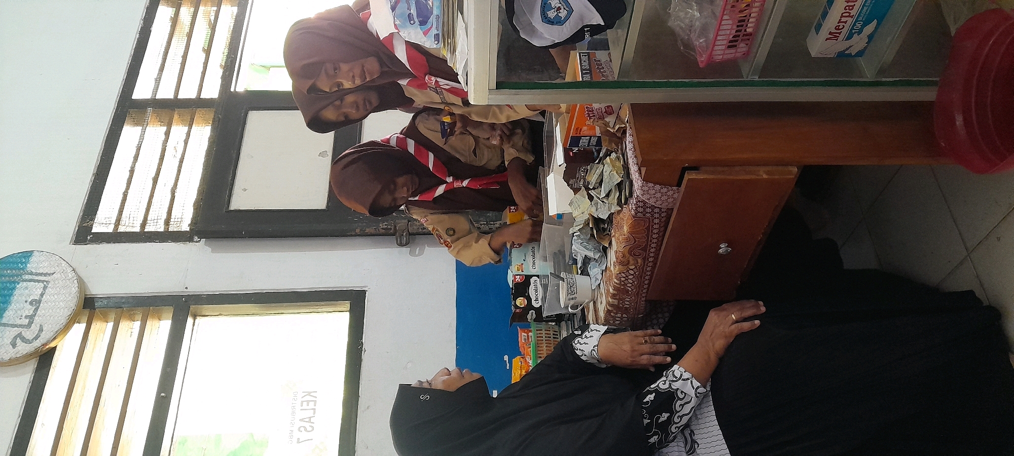 In Tahunan, Indonesia, a group of young girls in scout uniforms are gathered around a table filled with items, possibly in a classroom or shop. Two girls in the foreground are wearing brown scout uniforms and headscarves. One is looking down, while the other is looking towards the table. Behind them, another girl in a similar uniform is also looking down. A woman, also wearing a headscarf, stands to the right, her hands on the table. The table is covered with a patterned cloth and various items including boxes, bags, and what appear to be some coins or bills. In the background, there's a display shelf with products, including a box labeled "Merpati" and a red basket. A window with bars is visible on the left, and another window behind the woman on the right. The scene appears to be indoors, with tiled flooring.
