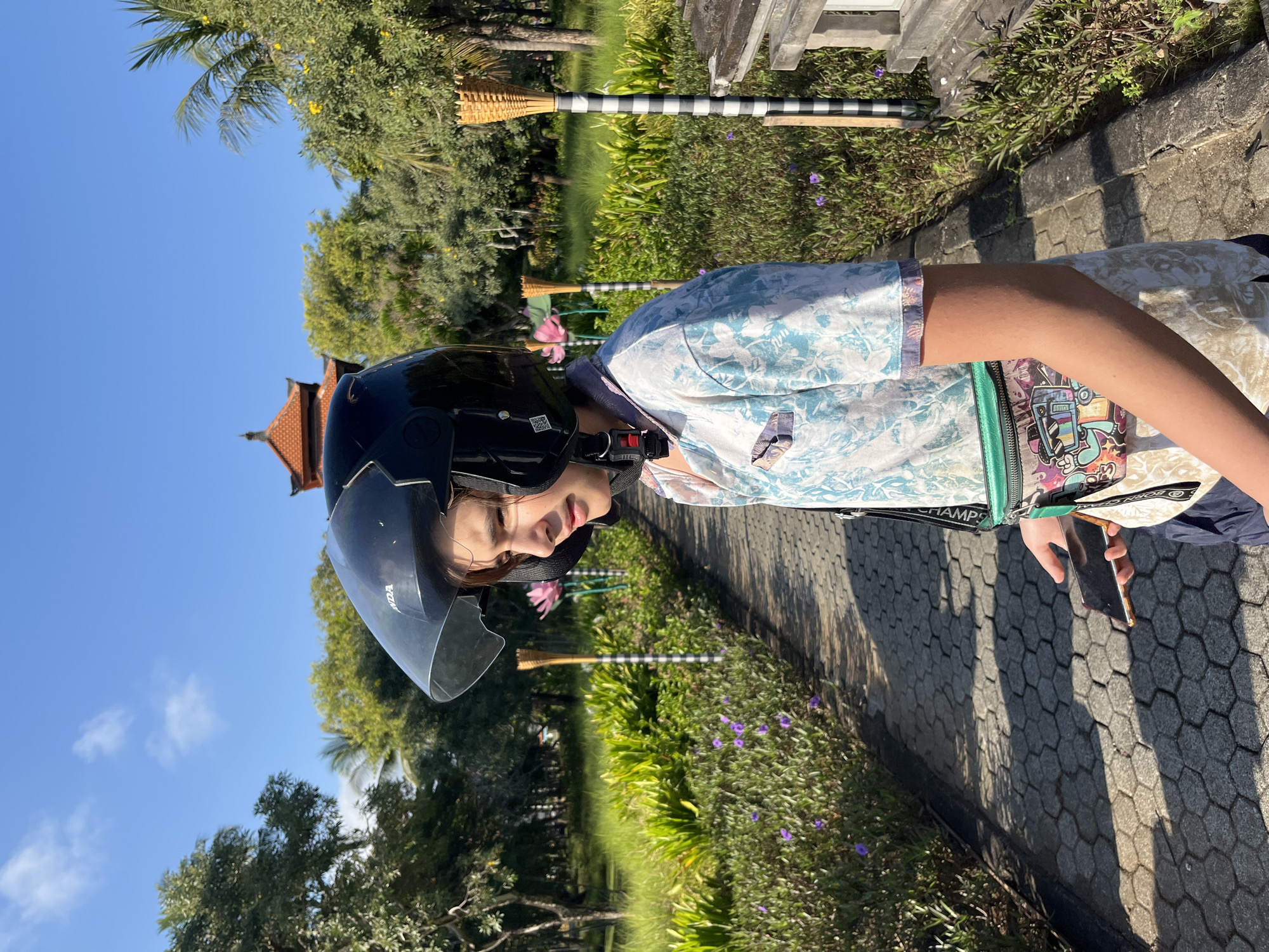 A young person wearing a black motorcycle helmet stands on a paved walkway in a garden area. The person is looking to the right and is wearing a light blue and white patterned short-sleeved shirt, a green fanny pack with colorful graphics, and dark blue shorts. They are holding a dark-colored smartphone in their right hand.
The walkway is made of hexagonal paving stones and is bordered by lush green foliage with scattered small purple flowers. In the background, there are trees, a traditional Balinese building with an orange tiled roof, and decorative torches with black and white striped poles. The sky is clear and blue with a few wispy clouds. The scene appears to be outdoors, likely in a resort or park setting in Kuta Selatan, Indonesia, given the architectural style and tropical vegetation.