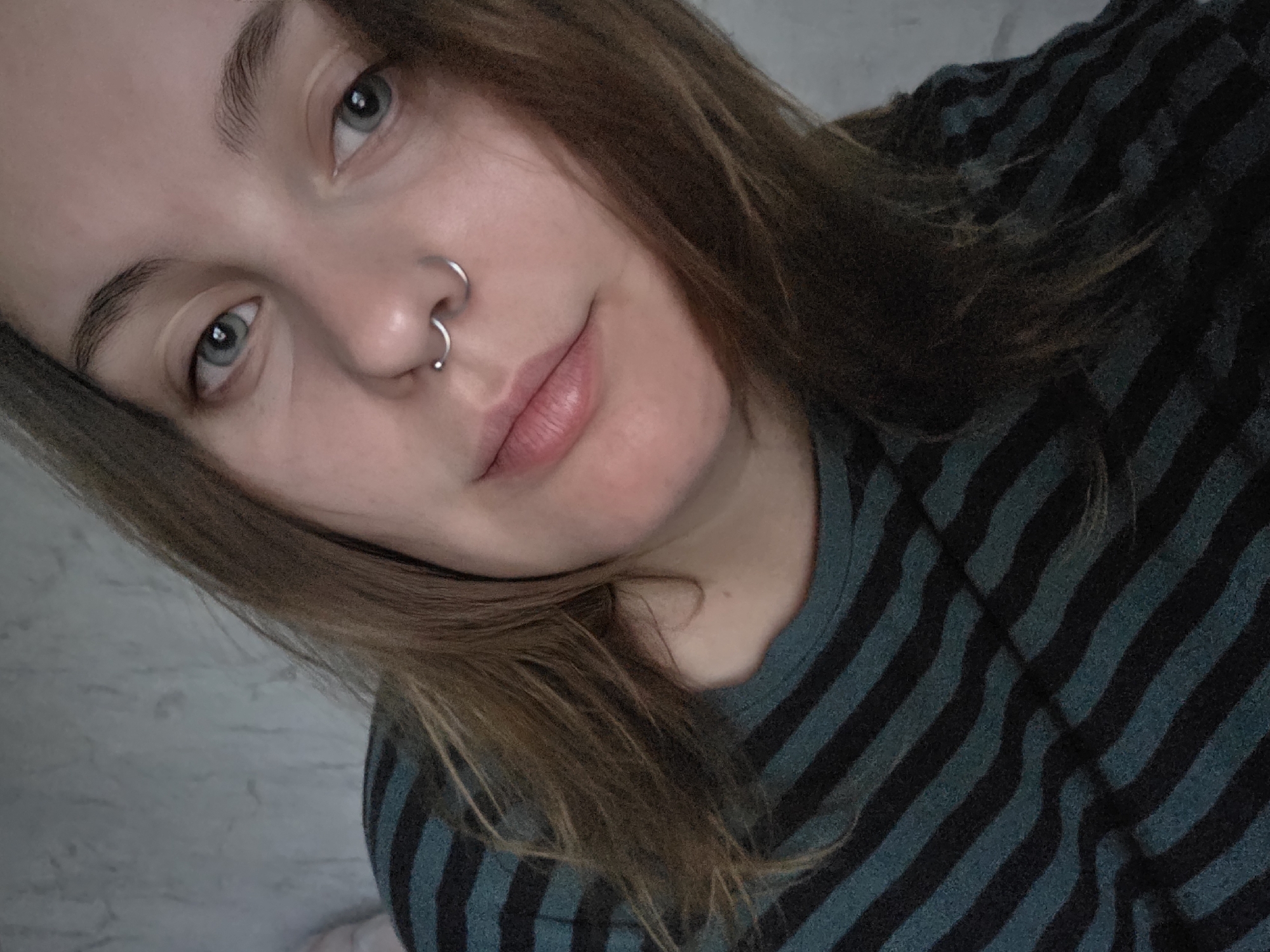 The image is a close-up selfie of a young woman with light blue eyes and medium brown hair. She is wearing a dark green and black striped shirt and has a septum piercing. The background appears to be a textured wall. The lighting is soft and diffused. There is no discernible location in the city of Minsk, Belarus.