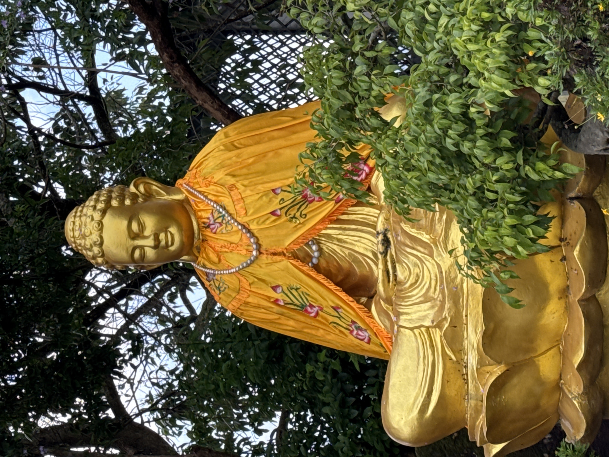 A golden Buddha statue sits in meditation on a lotus pedestal, adorned with a yellow robe featuring floral embroidery and a pearl necklace. The statue is partially obscured by lush green foliage and tree branches, with a glimpse of a lattice fence in the background. The scene is set outdoors, possibly in a garden or temple complex, within Lâm Viên - Đà Lạt, Vietnam. The lighting suggests it is daytime, with diffused light filtering through the trees. There are no people visible in the image.