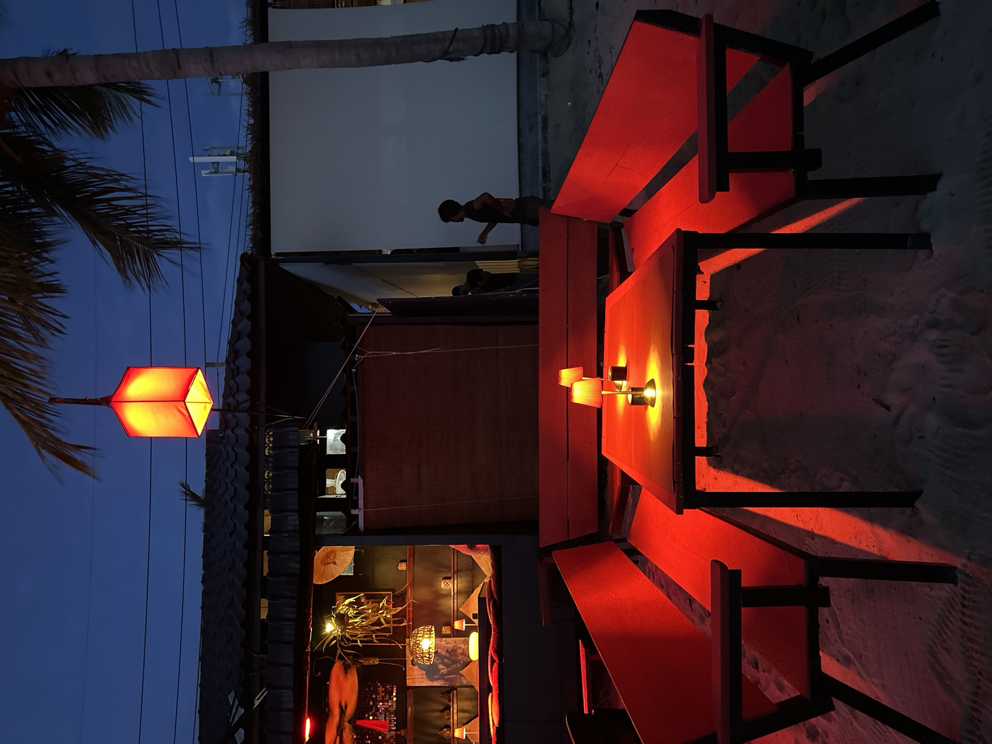 This media file depicts a scene at dusk or night in a beachside restaurant or bar, possibly in Ko Pha-ngan, Thailand. The dominant color is a vibrant red, cast by illuminated wooden tables and benches. A single, octagonal red lantern hangs from a wire to the left, casting an orange glow. Behind the seating area, a building with a dark roof and a lighter-colored wall is visible.
A silhouetted figure of a person can be seen standing near the building, appearing to be looking down or working. The ground is sandy, suggesting a beach location. Several small, yellow lamps are placed on the table, also contributing to the warm lighting. The sky is a deep blue, indicating it's twilight or nighttime. There are some electrical wires visible against the sky, and a palm tree frond extends from the left side of the frame. The overall mood is atmospheric and inviting, with the red lighting creating a cozy and perhaps romantic ambiance. No discernible text is visible in the image.