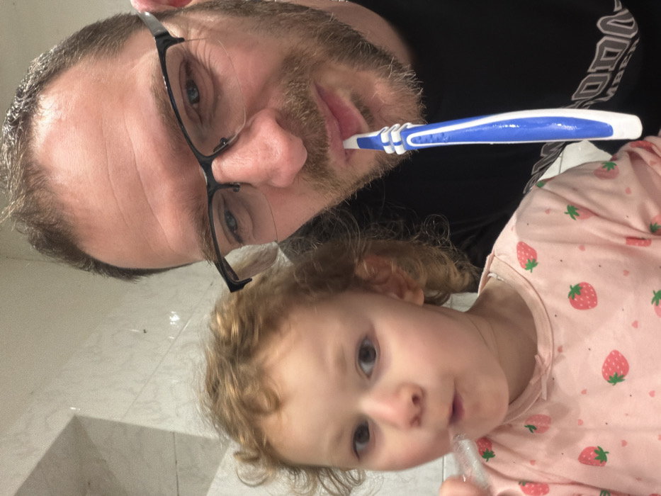 The image depicts an adult male and a young child inside what appears to be a bathroom located in Trassem, Germany. Both individuals are engaged in brushing their teeth. The male, positioned on the right, is light-skinned, has short brown hair, a stubble beard, and wears black-framed eyeglasses; he holds a blue and white toothbrush in his mouth. His visible right eye is blue. The child, positioned on the left, is light-skinned with curly reddish-blonde hair and wears a light pink long-sleeved shirt patterned with red strawberries; she holds a transparent toothbrush with white bristles in her mouth. The background consists of light-colored, possibly tiled, walls.
