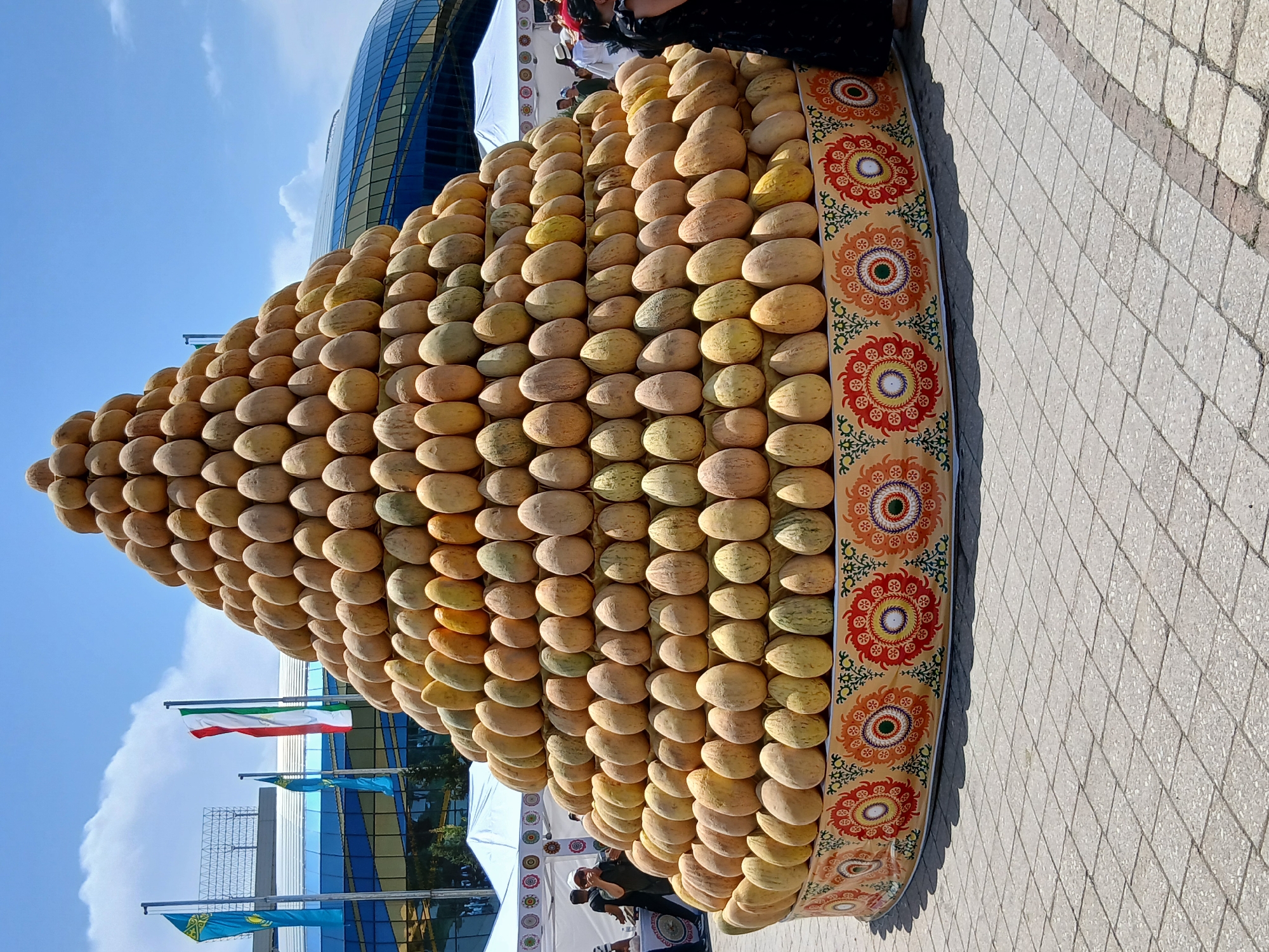 This image captures an outdoor scene featuring a large, pyramid-shaped display of melons. The melons are stacked in a visually appealing manner, forming a towering structure that is decorated with a brightly colored, patterned fabric along its base. The fabric features intricate floral motifs in red, orange, yellow, and green.
In the background, a modern building with a curved, glass facade and a blue tint rises against a clear blue sky with scattered clouds. Several national flags are visible, including one with red, white, and green stripes, and two blue flags with gold emblems, suggesting a possible cultural or national event.
At the top of the melon pyramid, several people are visible, some of whom appear to be spectators or participants. One person is wearing a dark headscarf and dark clothing. Further down, near the base of the display, other individuals can be seen, including someone wearing a white cap and a dark shirt. The overall impression is that of a festive occasion, perhaps a harvest festival, market, or cultural exhibition. The lighting suggests it is daytime, possibly late morning or early afternoon, with bright sunlight casting shadows on the paved ground. The ground itself is made of interlocking square paving stones. There is no visible text in the image.
