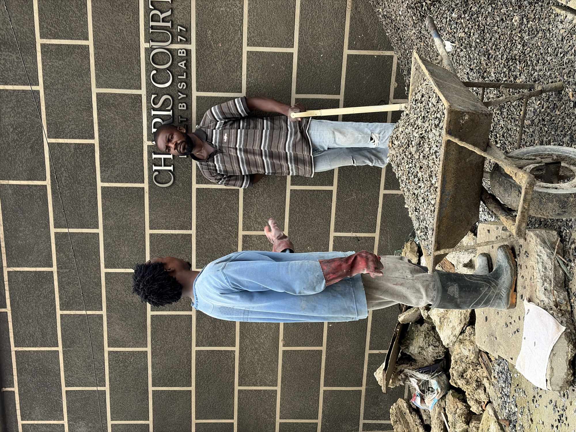 Two individuals are engaged in construction work in an outdoor setting. One man, wearing a striped t-shirt and distressed jeans, stands near a wall with the inscription "CHRIS COURT BY SLAB 77". He holds a wooden pole, possibly for construction purposes. Beside him, a wheelbarrow filled with gravel is partially visible. The other man, wearing a light blue long-sleeved shirt, light-colored pants, and red gloves, stands with his back to the camera. He appears to be wearing black rubber boots. The background features a dark brick wall and an abundance of gravel and construction debris. The scene suggests a daytime activity, likely related to building or renovation. The overall environment appears to be a construction site.