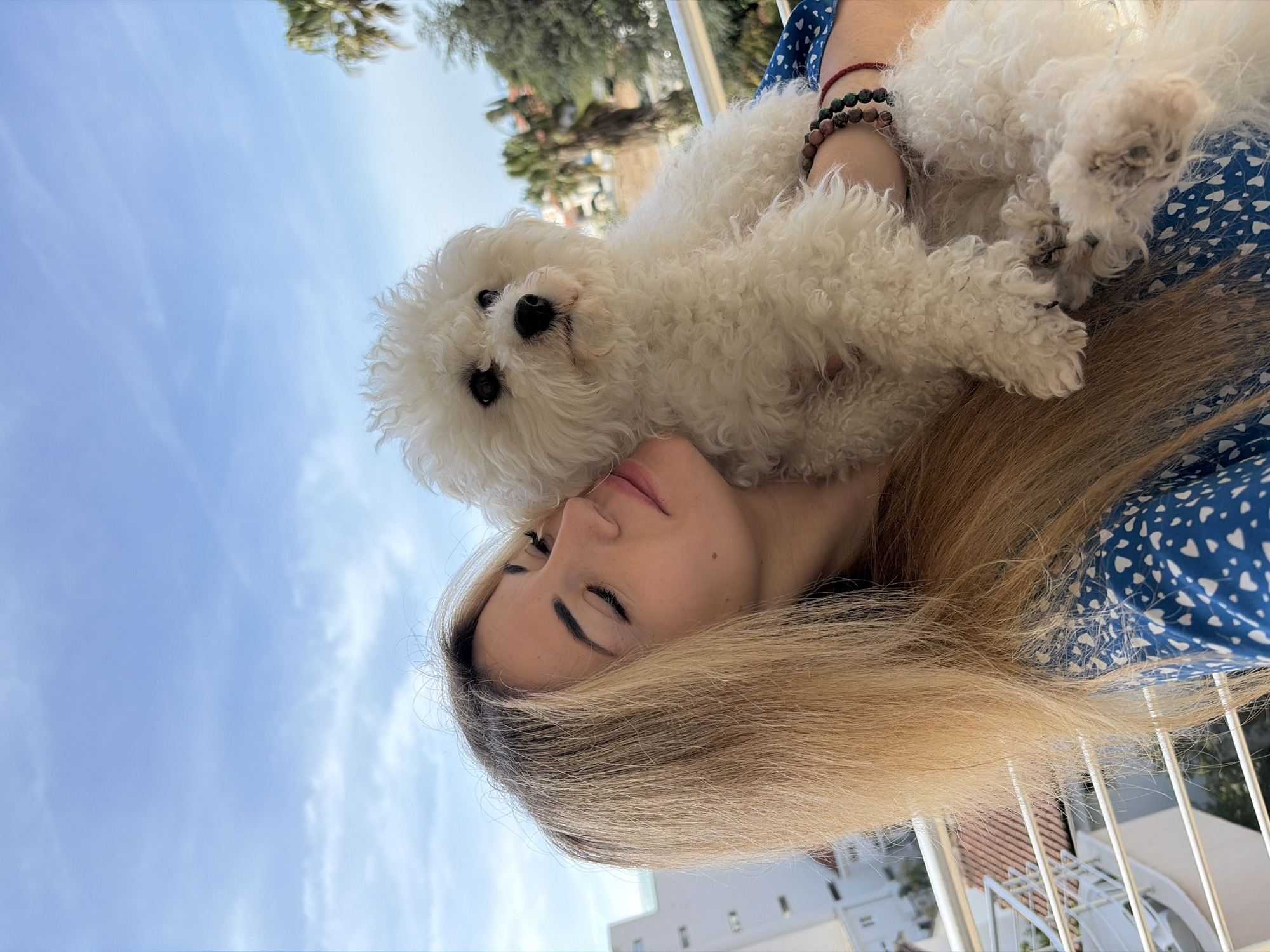 A woman with blonde hair is holding a white Bichon Frise dog close to her face. She is wearing a blue dress with a white heart pattern. The dog is resting on her shoulder and head. The background shows a clear blue sky with some trees and buildings. The scene appears to be taken outdoors, possibly on a balcony or rooftop. The location is possibly in Budva, Montenegro.