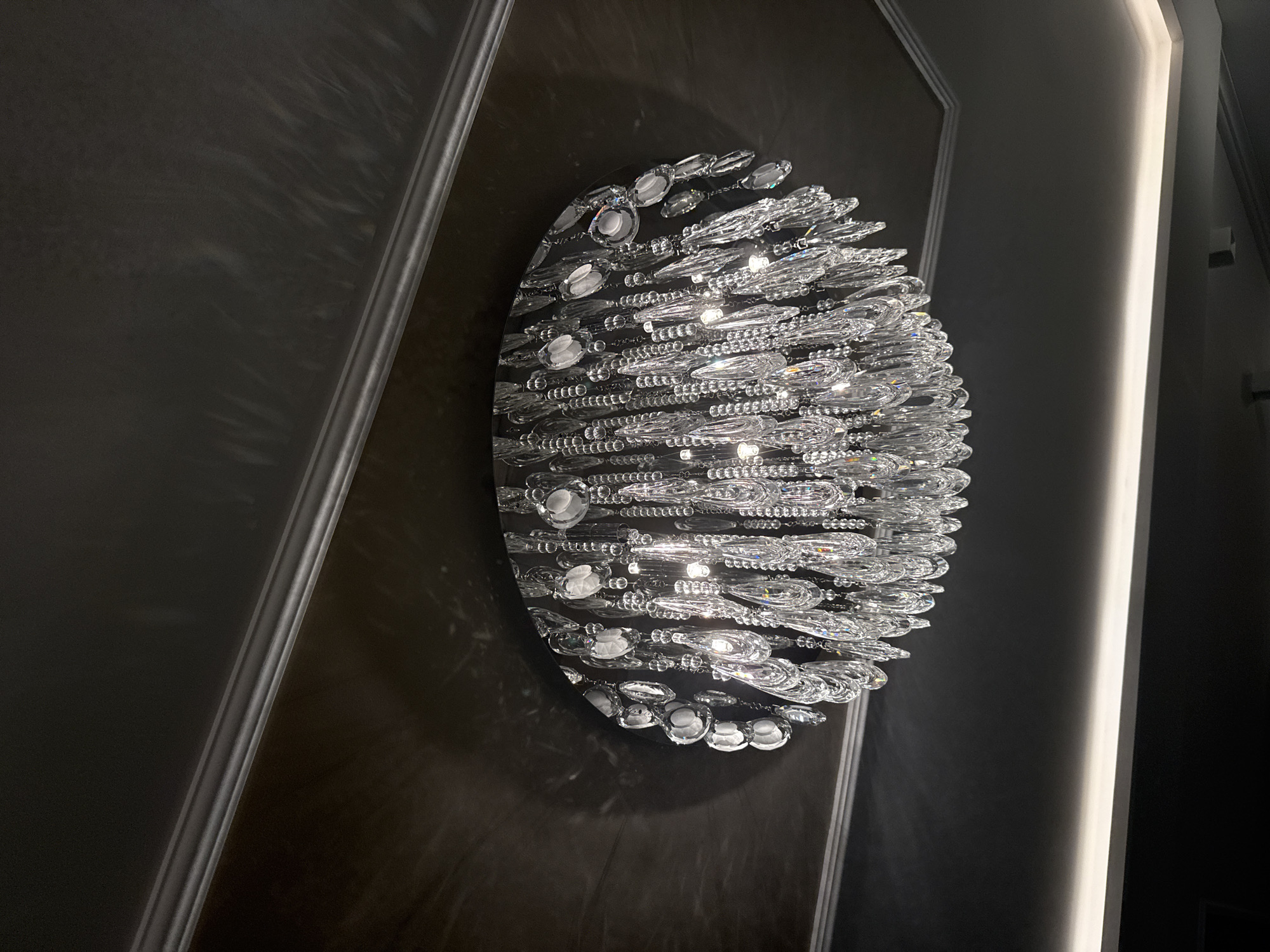 The image captures a close-up view of an ornate, oval-shaped chandelier mounted on a dark, possibly wood-paneled wall. The chandelier is predominantly clear, featuring numerous crystal or glass elements arranged in multiple layers, giving it a textured and sparkling appearance.
To the right of the chandelier, there is a vertical, bright white light strip, possibly an architectural lighting feature, which contrasts with the dark wall behind it. The wall itself seems to be dark gray or black with decorative molding.
The scene suggests an interior setting, possibly a hotel lobby, a high-end residential space, or a commercial establishment with sophisticated decor. There is no direct indication that the location is Sofia, Bulgaria within the image.