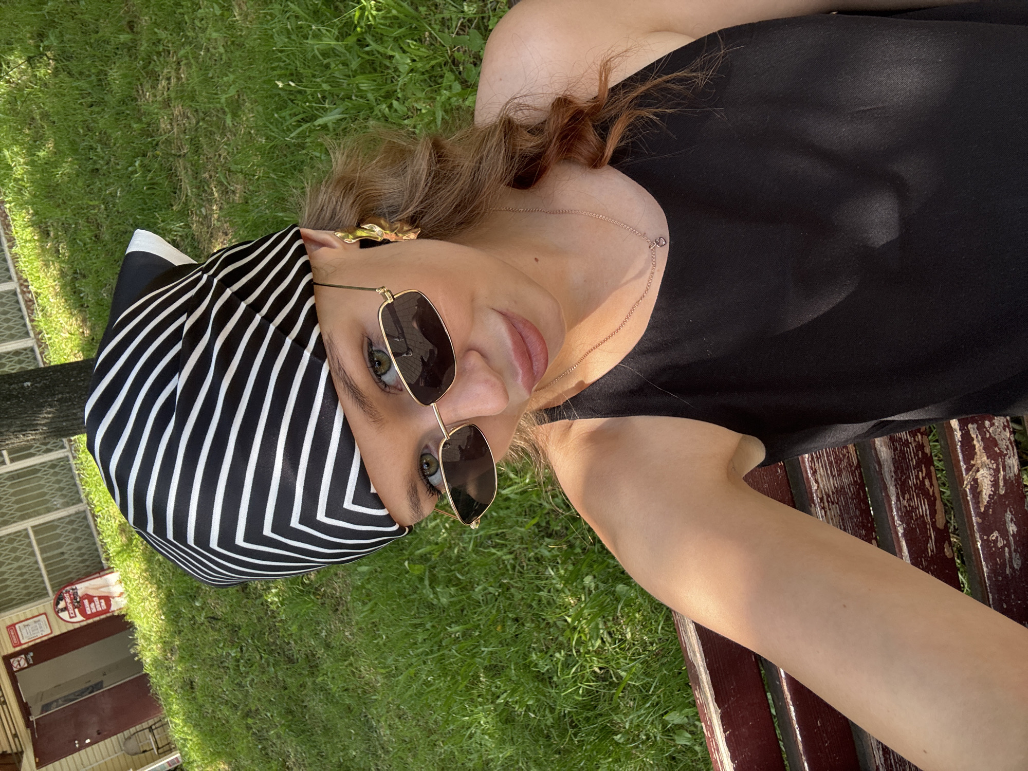 A young woman is lying on her back on a wooden bench outdoors. She is wearing a black top, sunglasses, and a black and white striped headscarf. Her hair is light brown and curly. The woman is looking up at the camera with a slight smile. The background shows green grass and trees. The scene appears to be set in a park or backyard on a sunny day. There is a building with red doors and windows visible in the background, with some signage. The overall mood is relaxed and casual. The text visible on the sign is "Russian Railways" and "O.K."