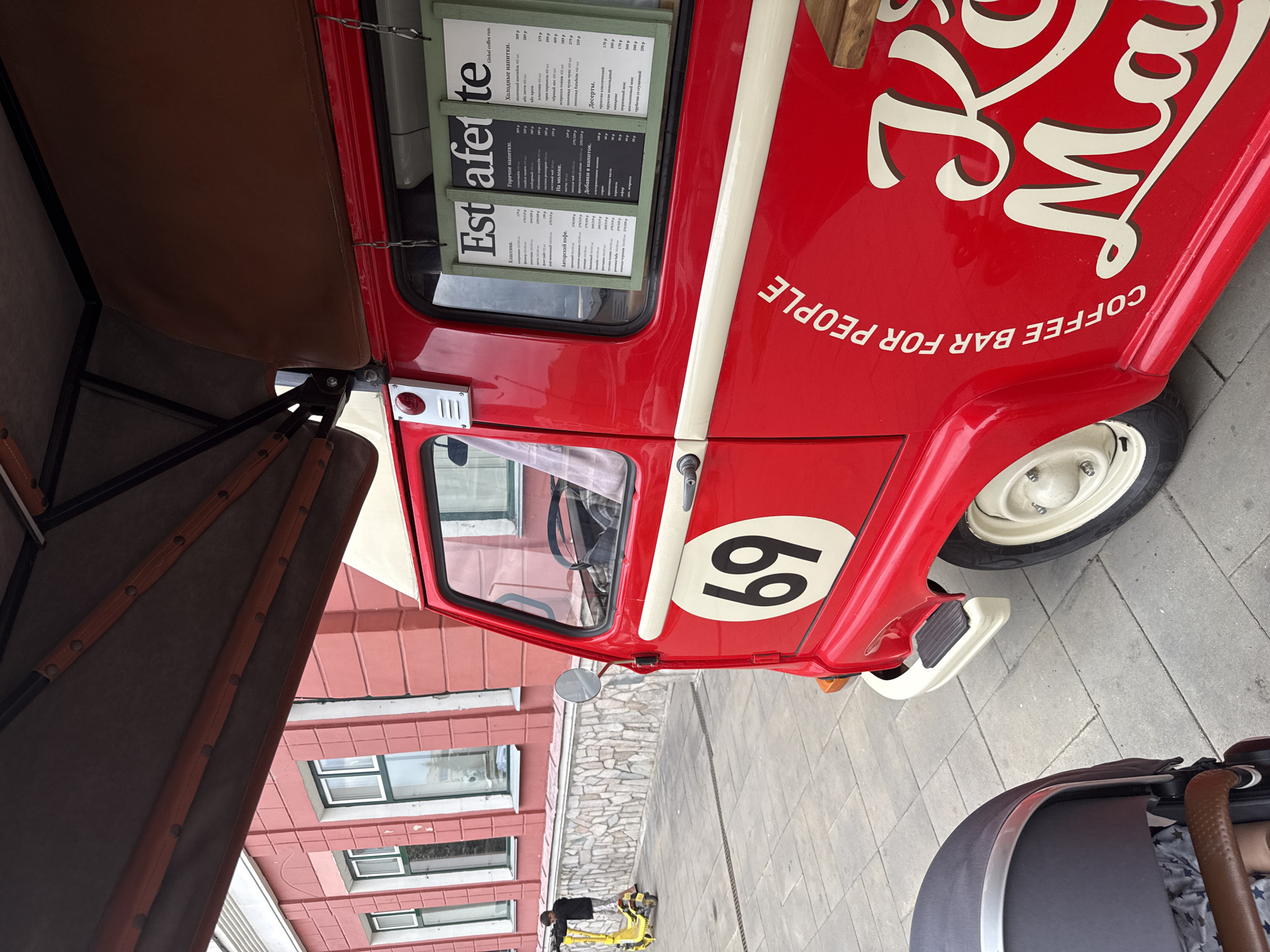 The image depicts a red food truck with a menu board hanging in the window. The menu is in Russian and lists various coffee drinks and desserts. A number "69" is visible on the side of the truck in a white circle. The truck is parked on a paved sidewalk next to a pink building with windows. A child on a yellow scooter is visible in the background. In the foreground, a stroller is partially visible. The scene appears to be a city street. The food truck is identified as "Kofemania", a Russian coffee shop chain, and is often found in urban locations. The presence of a stroller and a child on a scooter suggests a public area frequented by families. The food truck's design and branding are consistent with its operation as a mobile coffee bar. The overall atmosphere suggests a bright, daytime scene in a public urban setting.