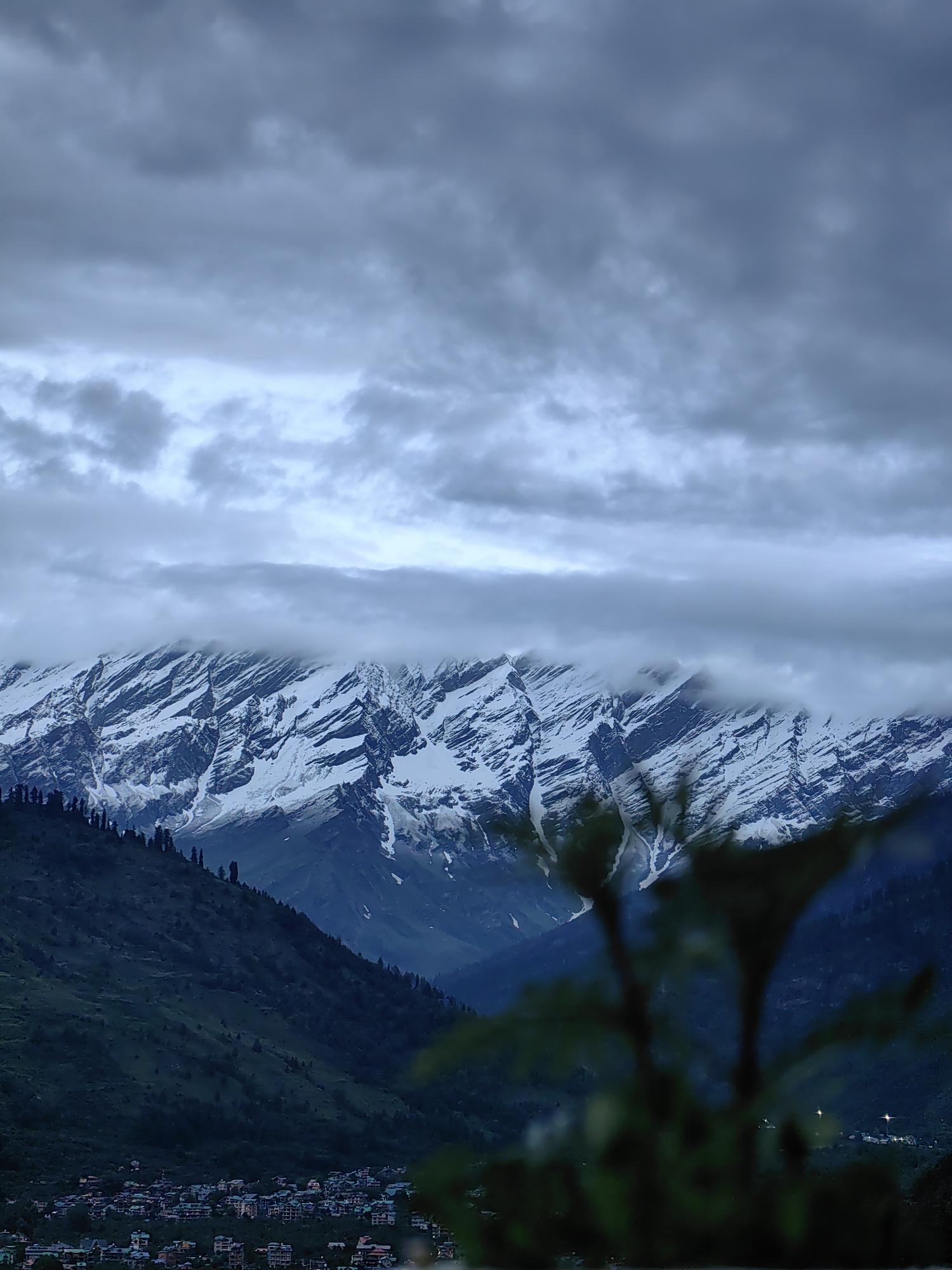 The image captures a wide shot of a mountainous landscape in Vashisht, India, on an overcast day. The foreground is dominated by dark, out-of-focus foliage, obscuring parts of the scene. Behind the foliage, a dark green, heavily wooded mountain slope descends towards a settlement at its base. This settlement appears to be a town or village, characterized by numerous buildings with varied roof colors, clustered together. Further in the background, towering snow-capped mountains rise majestically. The peaks are jagged and striated with snow, with some areas obscured by a thick band of dark, low-hanging clouds that appear to be partially covering the mountains. Above this cloud layer, the sky is filled with more layers of lighter, diffused clouds, creating a somber and moody atmosphere. The overall impression is one of a remote, natural setting with a human settlement nestled at the foot of imposing, snow-clad mountains under a cloudy sky.