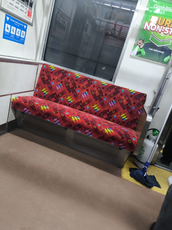 The image displays the interior of a commuter train, consistent with services operating in or around Cikarang, Indonesia. A two-person bench, upholstered in a red fabric with a repetitive pattern of multi-colored geometric shapes (rectangles and circles), is positioned along the left wall. The bench's base is a metallic structure with horizontal perforations.
The train interior features white wall panels. A large tinted window occupies the upper section of the wall, reflecting indistinct external structures. Above the window, two signs are mounted: a white rectangular sign with black text and symbols, and a blue square sign featuring four white pictograms (indicating priority seating for elderly, pregnant, disabled, and passengers with infants) and the Indonesian text "Tempat duduk prioritas."
To the right, a green and white GrabBike advertisement poster is affixed to the wall, depicting a person in a green jacket. Below the advertisement, on a yellow-striped section of the floor, a white pressurized tank with a green handle and an attached blue string mop head are leaning against the wall. The train floor in the foreground is a solid brown color. A person's lower leg, clad in dark trousers, and a white shoe are partially visible in the bottom right corner of the frame. The bench is unoccupied.