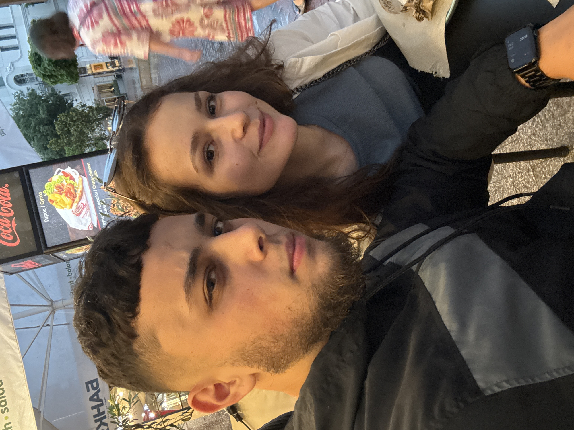 A slightly tilted, close-up portrait captures two young adults in an outdoor urban setting during the evening. The male subject is in the foreground, looking directly at the camera with a neutral, relaxed expression. He has short dark hair with a faded cut, a short beard, and is wearing a dark jacket, possibly black with grey accents. A black smartwatch with a metallic band is visible on his left wrist, displaying the time "20:11".
Behind and slightly to his right, the female subject looks towards the camera with a gentle smile. She has long, dark brown hair, with sunglasses resting on top of her head. She appears to be wearing a light blue or grey top under a white outer layer.
The background reveals a bustling street scene. To the left, a partially visible food stall sign features the "Coca-Cola" logo prominently, alongside text advertising "Gyros" and "Crispy chicken." Further back, blurred green trees and a white building with classical architectural elements are discernible. Several outdoor umbrellas provide shade, one bearing the Cyrillic word "БАНКА" (Bank). The ambient lighting, consistent with the smartwatch's time, suggests it is dusk or early night in Varna, Bulgaria. Both individuals appear relaxed, possibly enjoying a meal or a casual evening out.