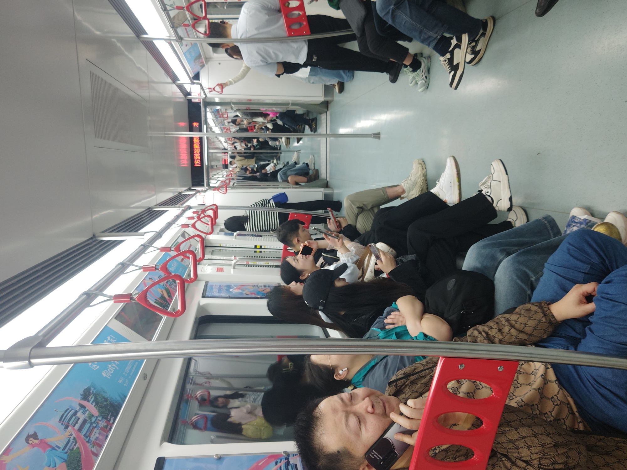 The interior of a subway car is shown in an image. Passengers are seated facing each other. Most of the passengers are looking at their phones. Some passengers are on the phone. The subway car has advertisements on the sides and red triangular handles hanging from the ceiling. The handles are suspended from a silver bar. The location is Unknown, Unknown.