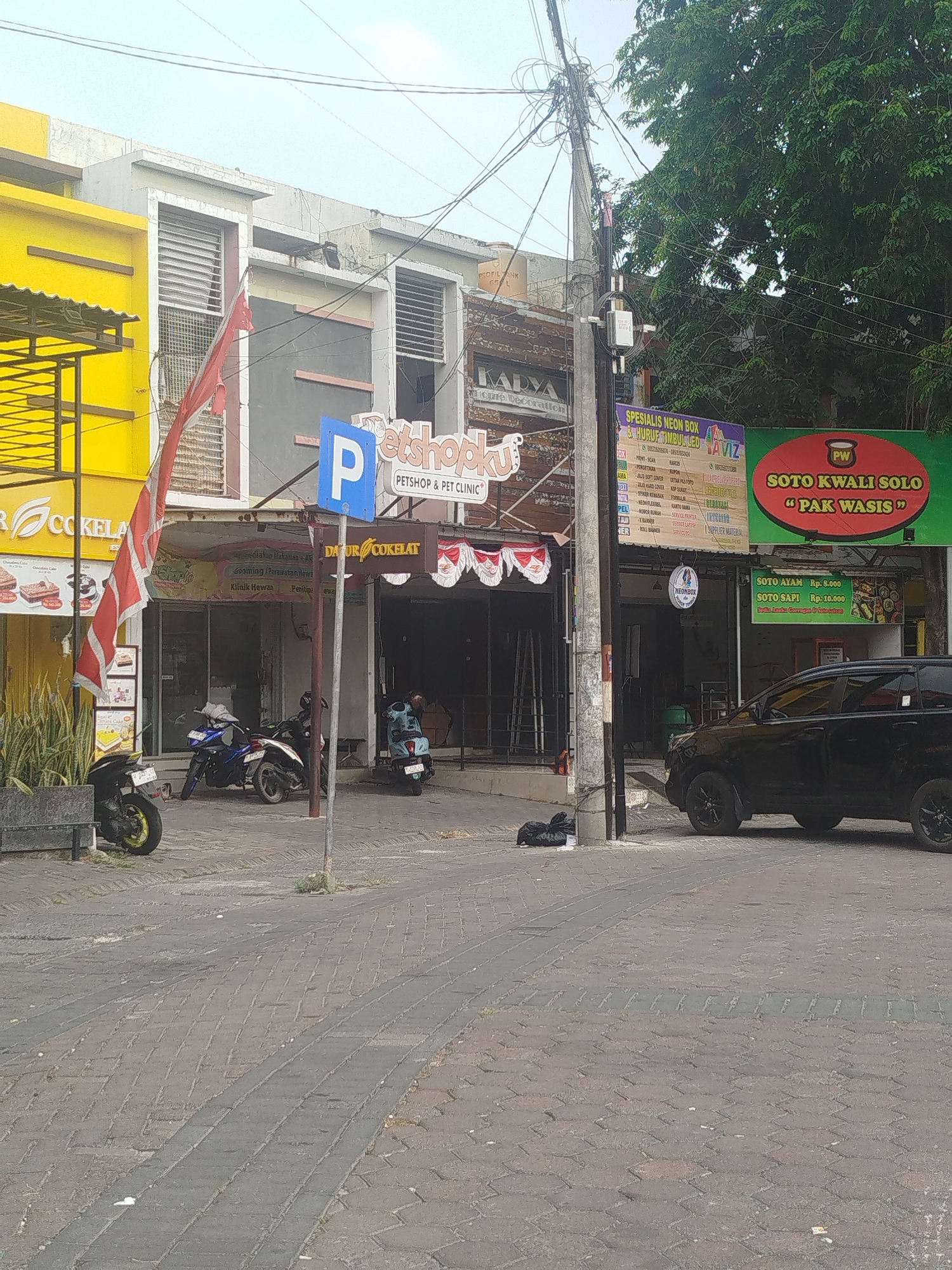 The image shows a street scene in Yosowilangun, Indonesia, featuring a row of shops and a street with brick paving. In the foreground, a black car is parked on the right side of the street. To the left, several motorcycles are parked. A prominent yellow building with a metal awning is visible on the far left, displaying a sign with "COKELAT" and images of cakes. Adjacent to it, a darker building has signs for "Petshopku PETSHOP & PET CLINIC" and "DATUR COKELAT". Further down the street, a green sign reads "SOTO KWALJ SOLO PAK WASIS". A utility pole with numerous wires is positioned in the middle of the scene. The environment appears to be a commercial area with outdoor seating or waiting areas outside the shops. The sky is overcast.